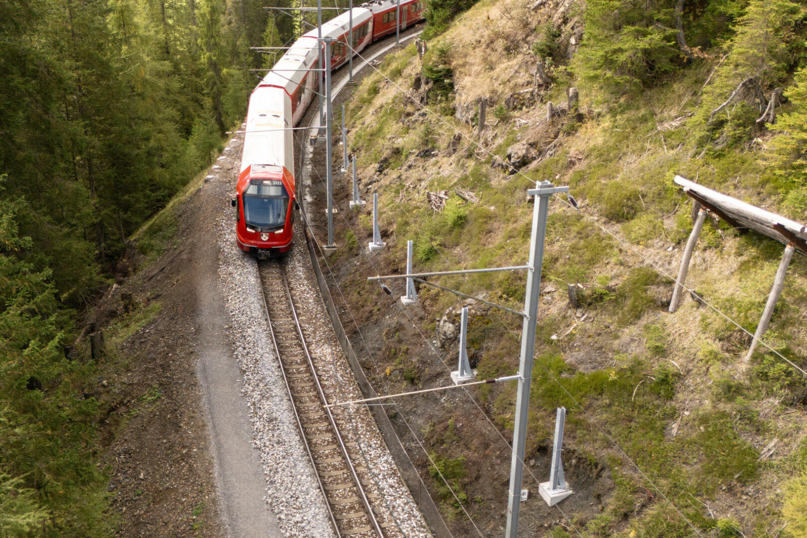 Ein roter Personenzug fährt auf einer gebogenen Bahnstrecke, umgeben von grünem Wald und steilen Grashängen, mit sichtbaren elektrischen Oberleitungen.