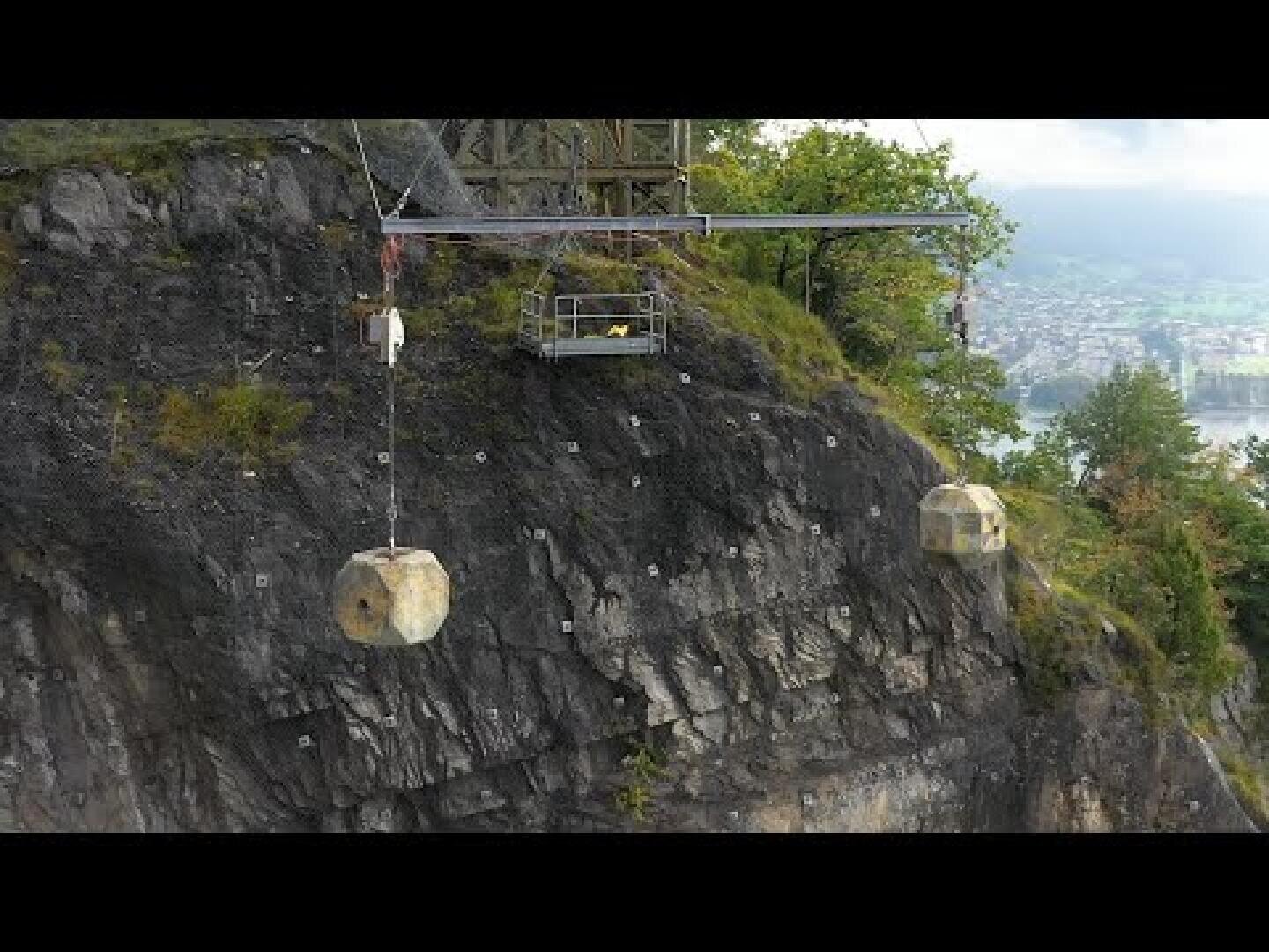 Ein steiler felsiger Abhang mit zwei großen Steinblöcken, die an Seilen aufgehängt sind, und einer Metallkonstruktion darüber, umgeben von Bäumen, mit Blick auf eine Stadt und Wasser im Hintergrund.