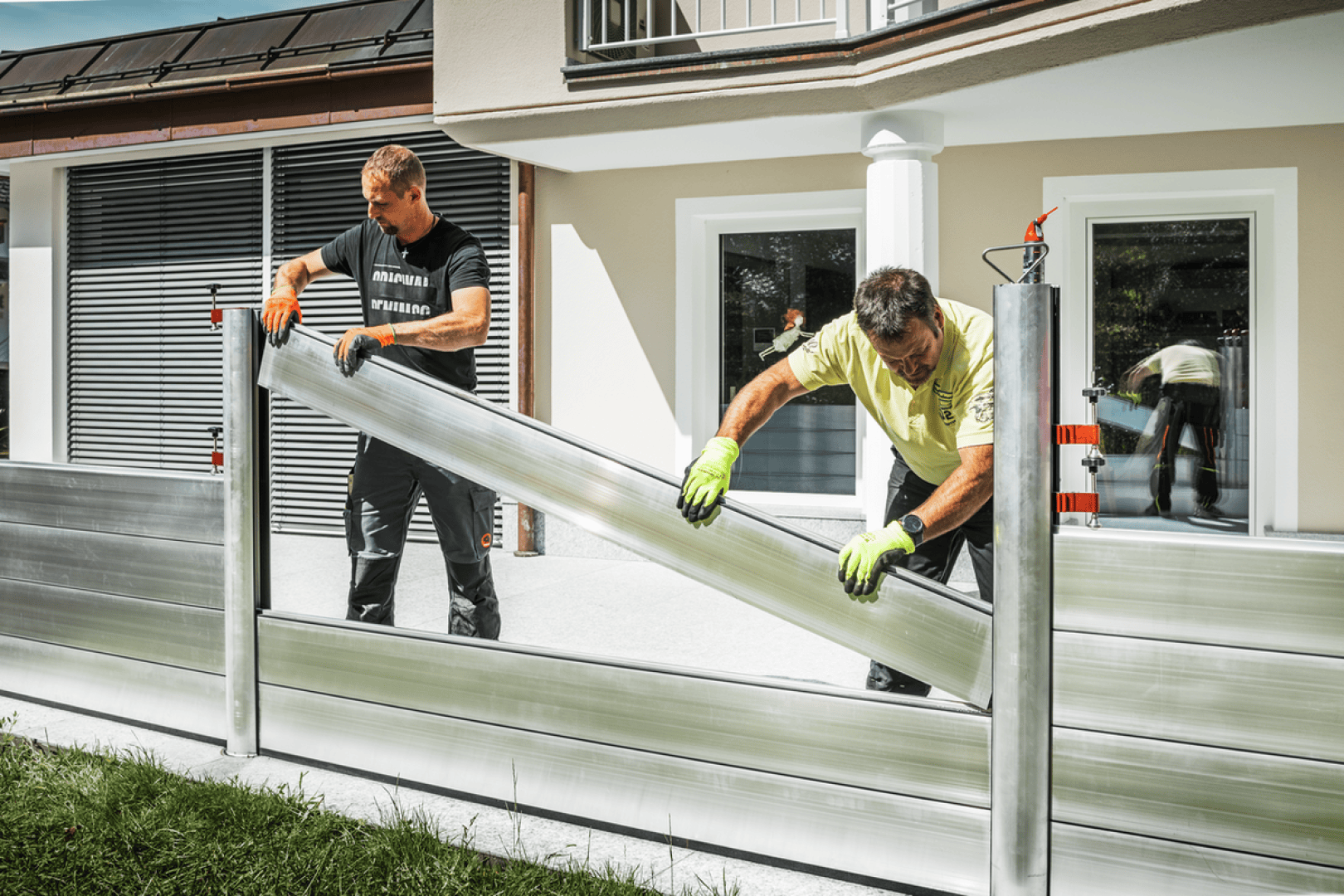 Zwei Männer mit Handschuhen montieren eine Hochwasserschutzwand aus Aluminium vor einem modernen beigen Haus mit Säulen und grossen Fenstern.