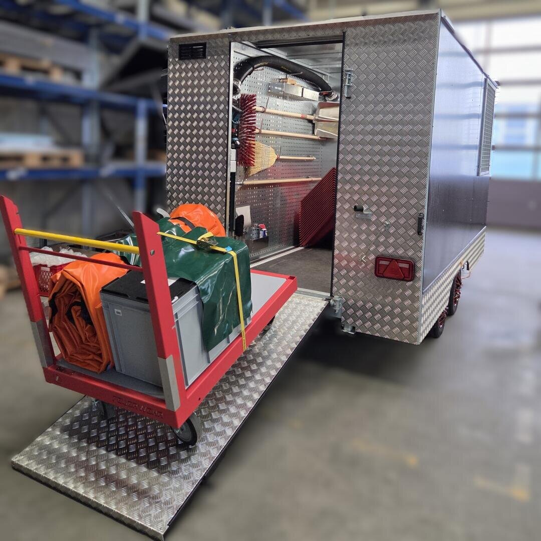 In einem Anhänger mit offener Hecktür und Rampe befinden sich Reinigungsmittel, Werkzeuge und Ausrüstung. Ein mit Behältern, einer Plane und einer orangefarbenen Jacke beladener Wagen steht am unteren Ende der Rampe.