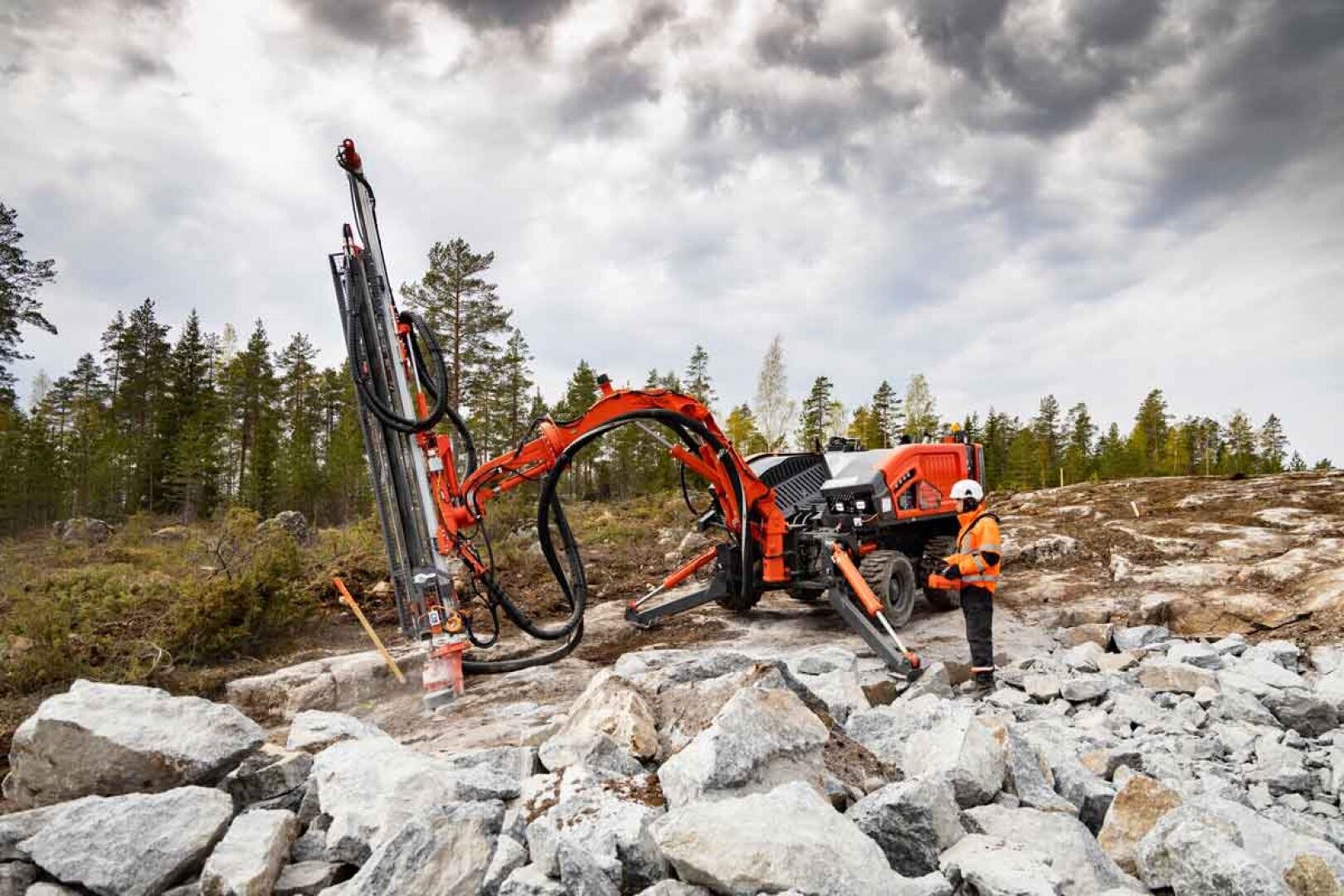 Ein Arbeiter in Schutzkleidung bedient eine große rote Felsbohrmaschine auf einer felsigen Baustelle mit verstreuten Felsbrocken im Freien, umgeben von Bäumen unter einem bewölkten Himmel.