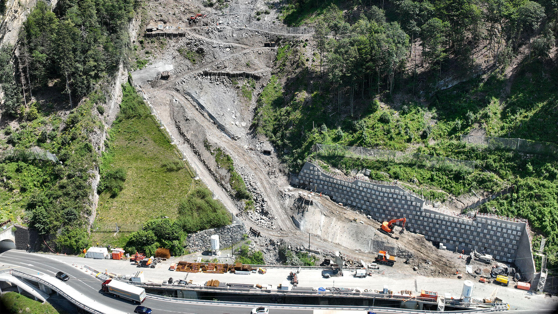 Luftaufnahme einer Baustelle an einem Hang in der Nähe einer Autobahn. Bagger und Baufahrzeuge arbeiten auf terrassiertem, gerodetem Land mit Stützmauern und freiliegendem Boden, umgeben von grünen Bäumen und Vegetation.