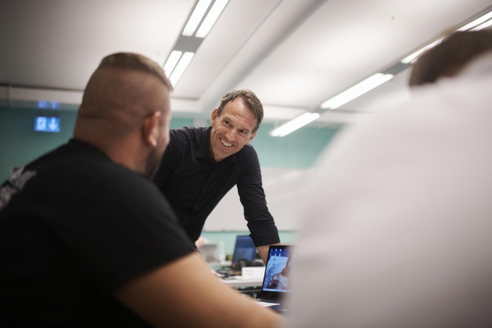 Ein Mann lächelt, während er sich über einen Tisch beugt und mit zwei Personen in einem hell erleuchteten Büro oder Klassenzimmer ein Gespräch führt. Auf dem Tisch im Vordergrund befindet sich ein Laptop.