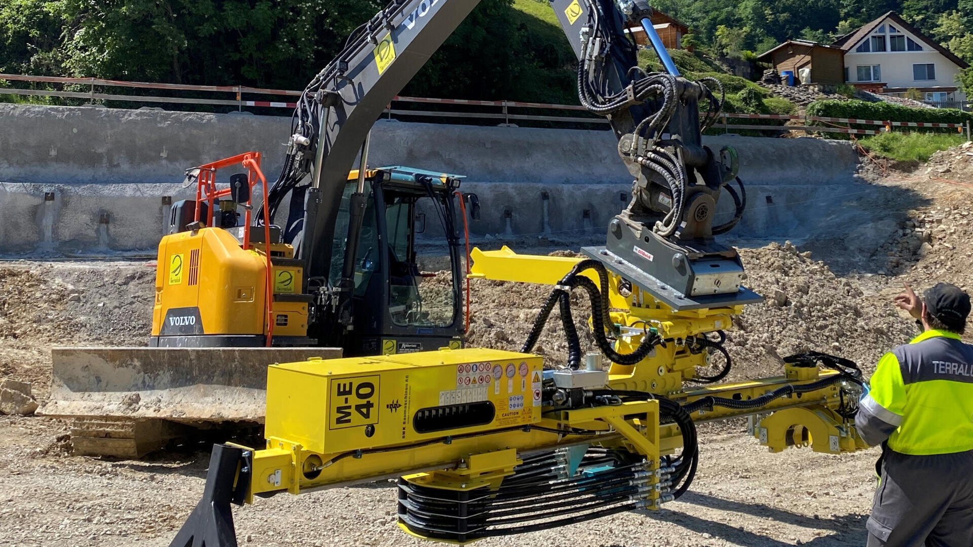 Ein Bauarbeiter steht neben einer gelben Bohrmaschine, die an einem Bagger auf einer Baustelle befestigt ist. Im Hintergrund sind eine Betonstützmauer und Häuser zu sehen.