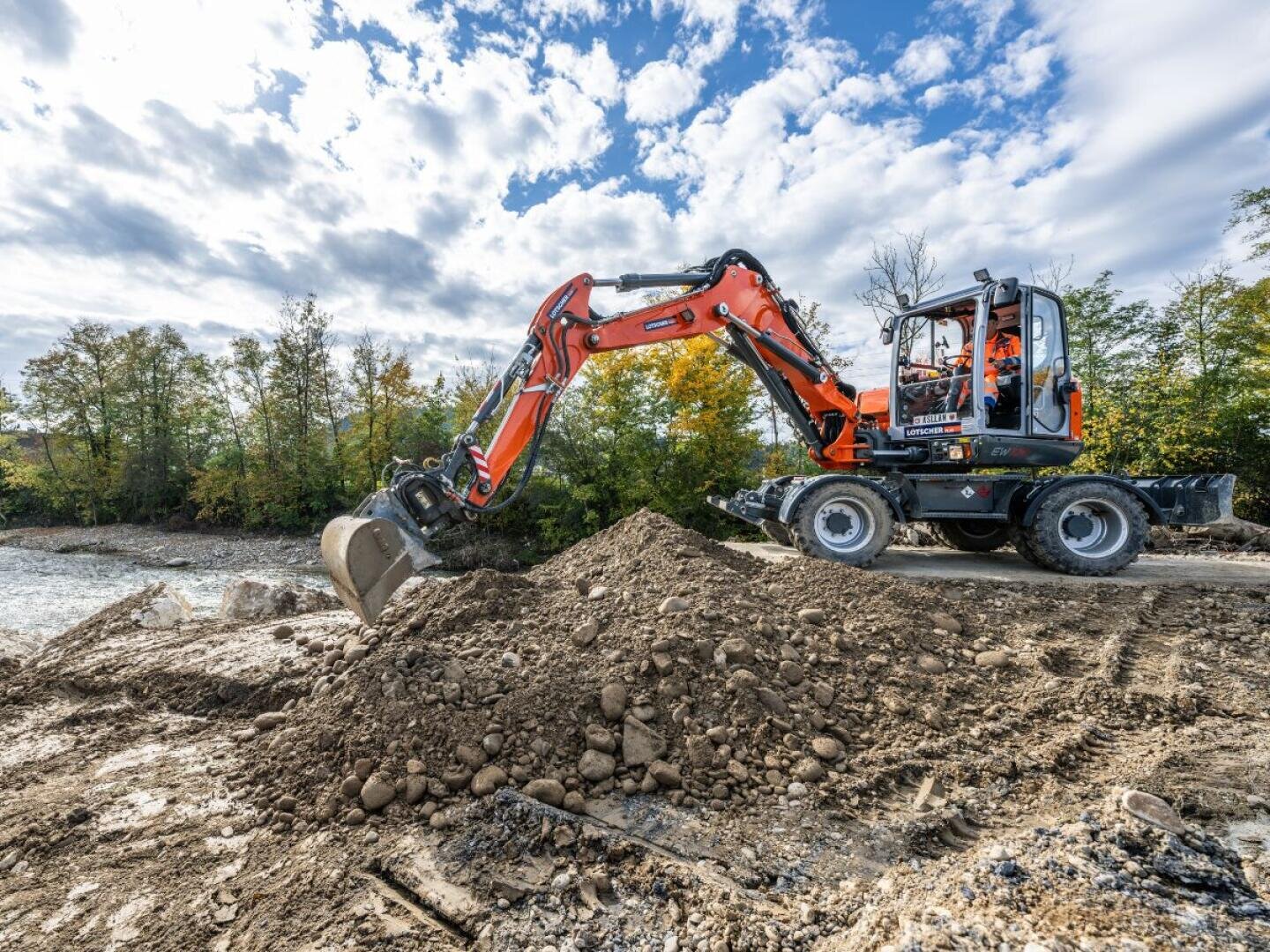 Ein orangefarbener Bagger bewegt einen Haufen Erde auf einer Baustelle, mit Bäumen und einem teilweise bewölkten Himmel im Hintergrund.