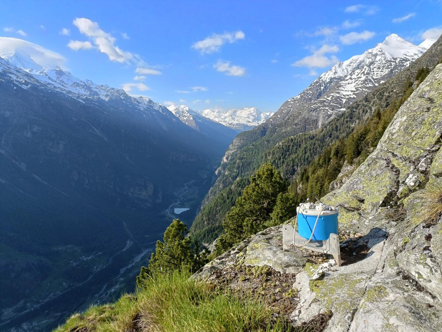 Ein blaues wissenschaftliches Instrument oder eine Wetterstation befindet sich auf einem mit Gras bewachsenen Felshang mit Blick auf ein tiefes Tal, umgeben von bewaldeten Bergen und schneebedeckten Gipfeln unter einem klaren blauen Himmel.