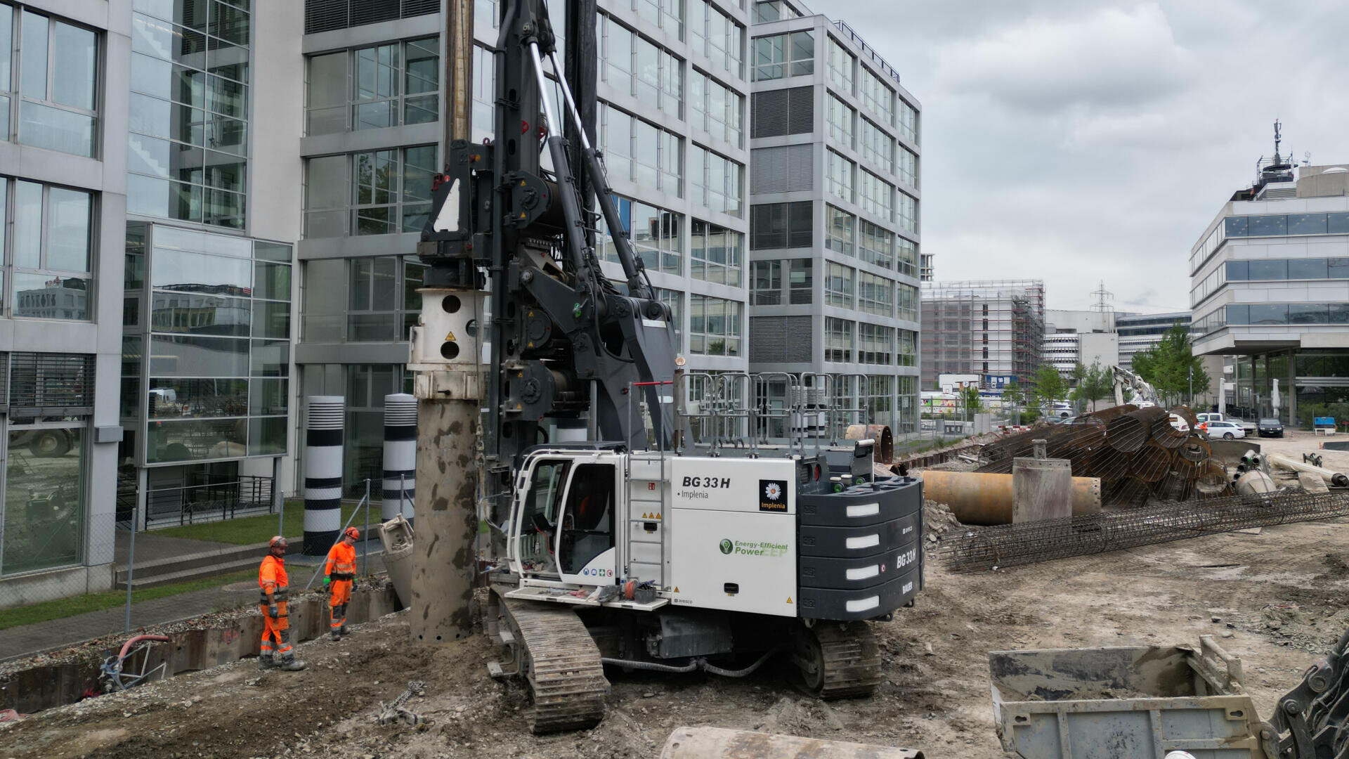 Eine große Bohrmaschine arbeitet auf einer Baustelle in einem städtischen Gebiet, umgeben von modernen Bürogebäuden. Zwei Arbeiter in orangefarbener Schutzkleidung stehen in der Nähe und beaufsichtigen die Arbeit.