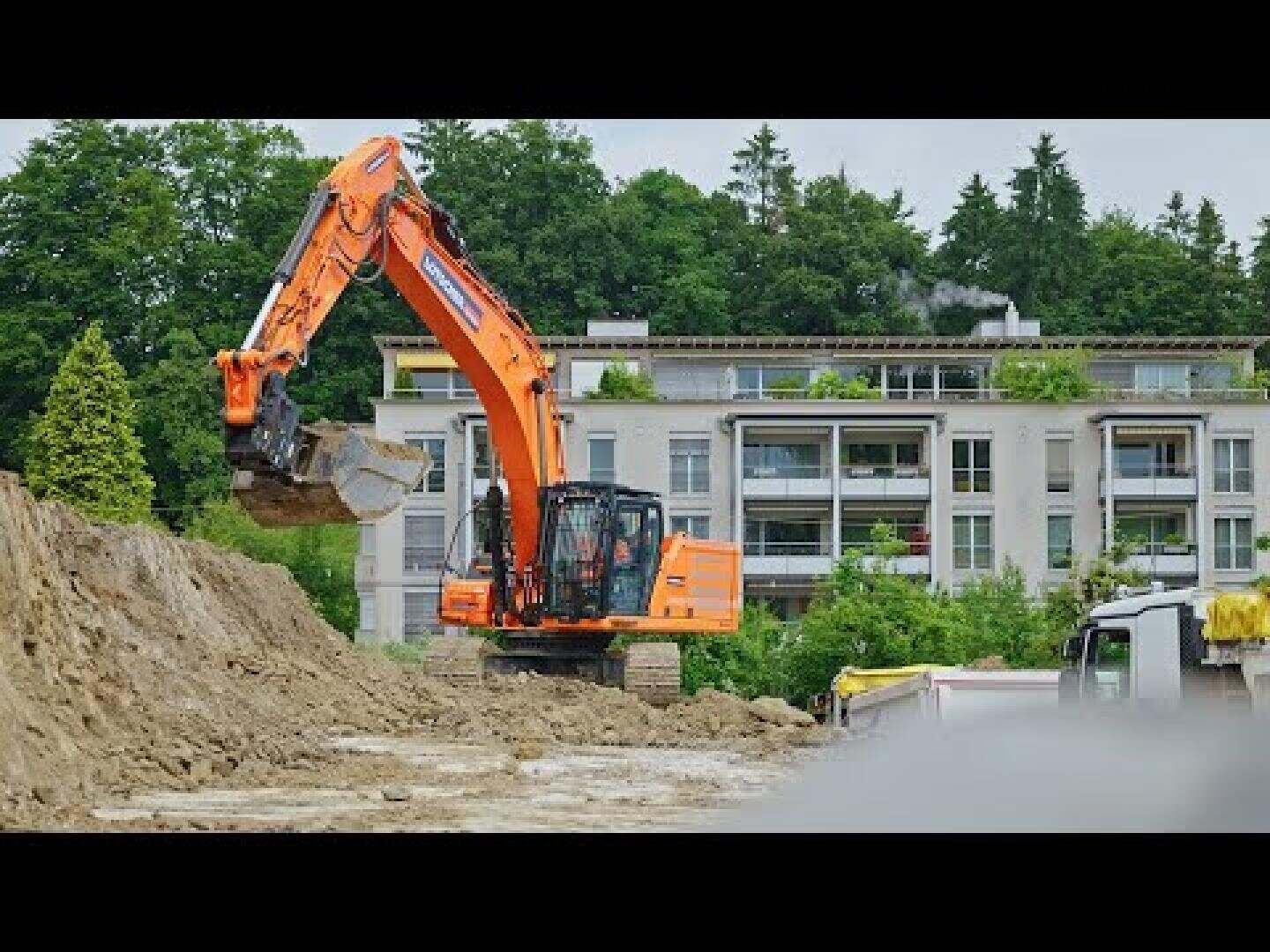 Ein orangefarbener Bagger schaufelt Erde auf einer Baustelle in der Nähe eines modernen Wohnhauses. Im Hintergrund sind grüne Bäume zu sehen, und rechts ist teilweise ein Lastwagen zu erkennen.