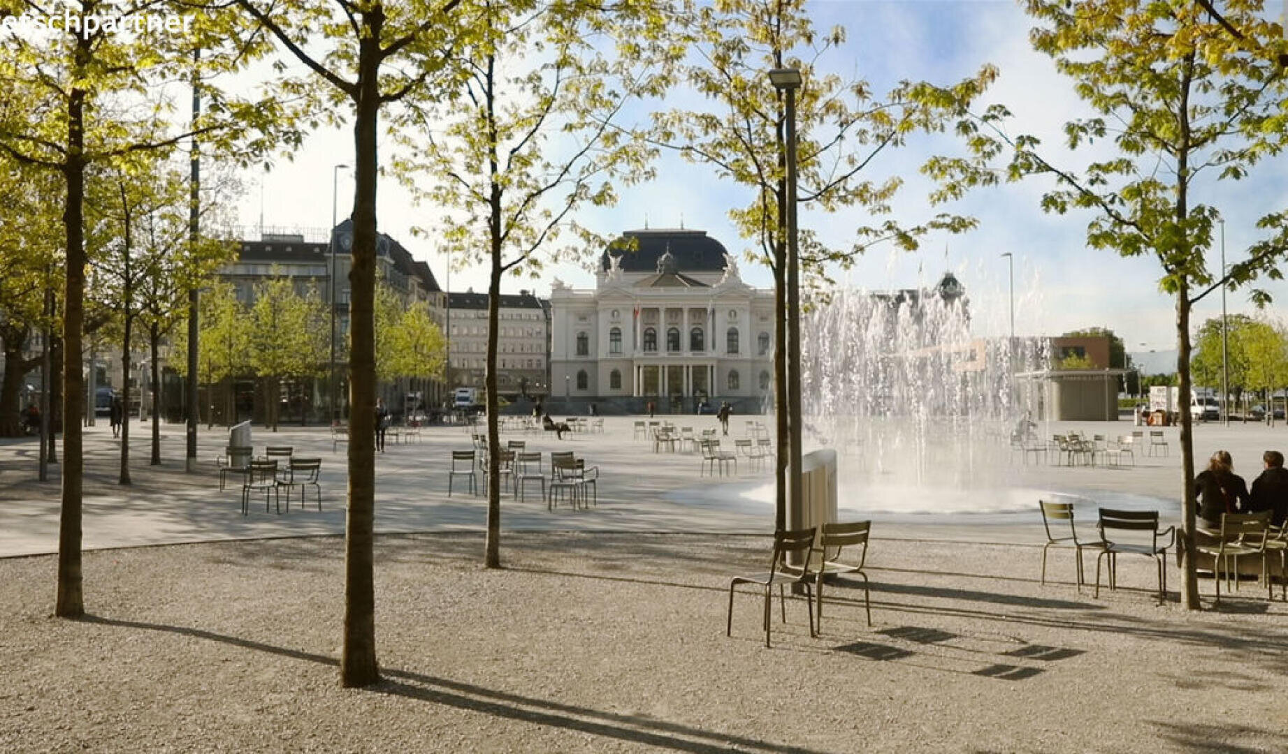 Ein sonniger Stadtplatz mit verstreuten Bäumen, Metallstühlen und sitzenden Menschen. In der Mitte sprudelt ein großer Springbrunnen, im Hintergrund ein großes historisches Gebäude und moderne Gebäude, die den Platz umgeben.