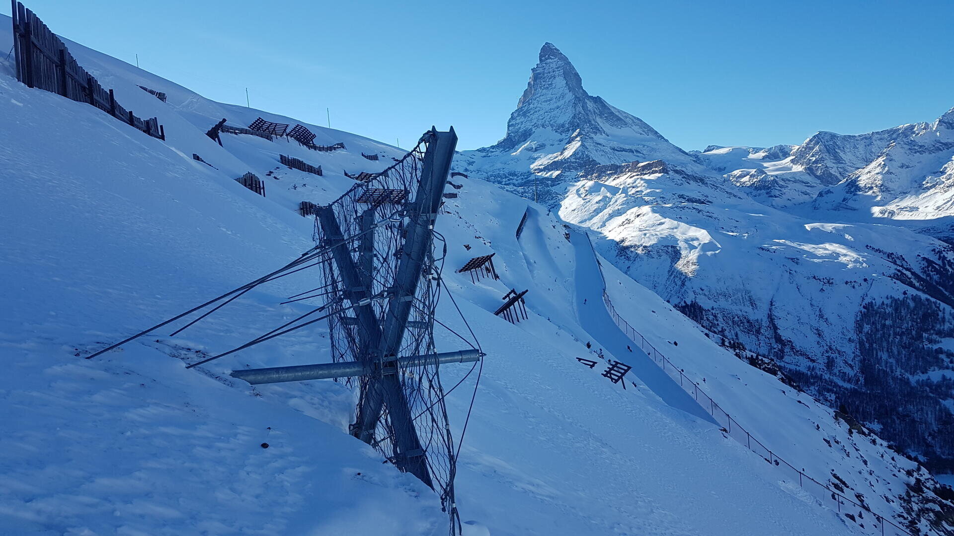 Lawinenverbauungen aus Metall stehen entlang eines schneebedeckten Hangs mit dem Matterhorn im Hintergrund unter einem klaren blauen Himmel.