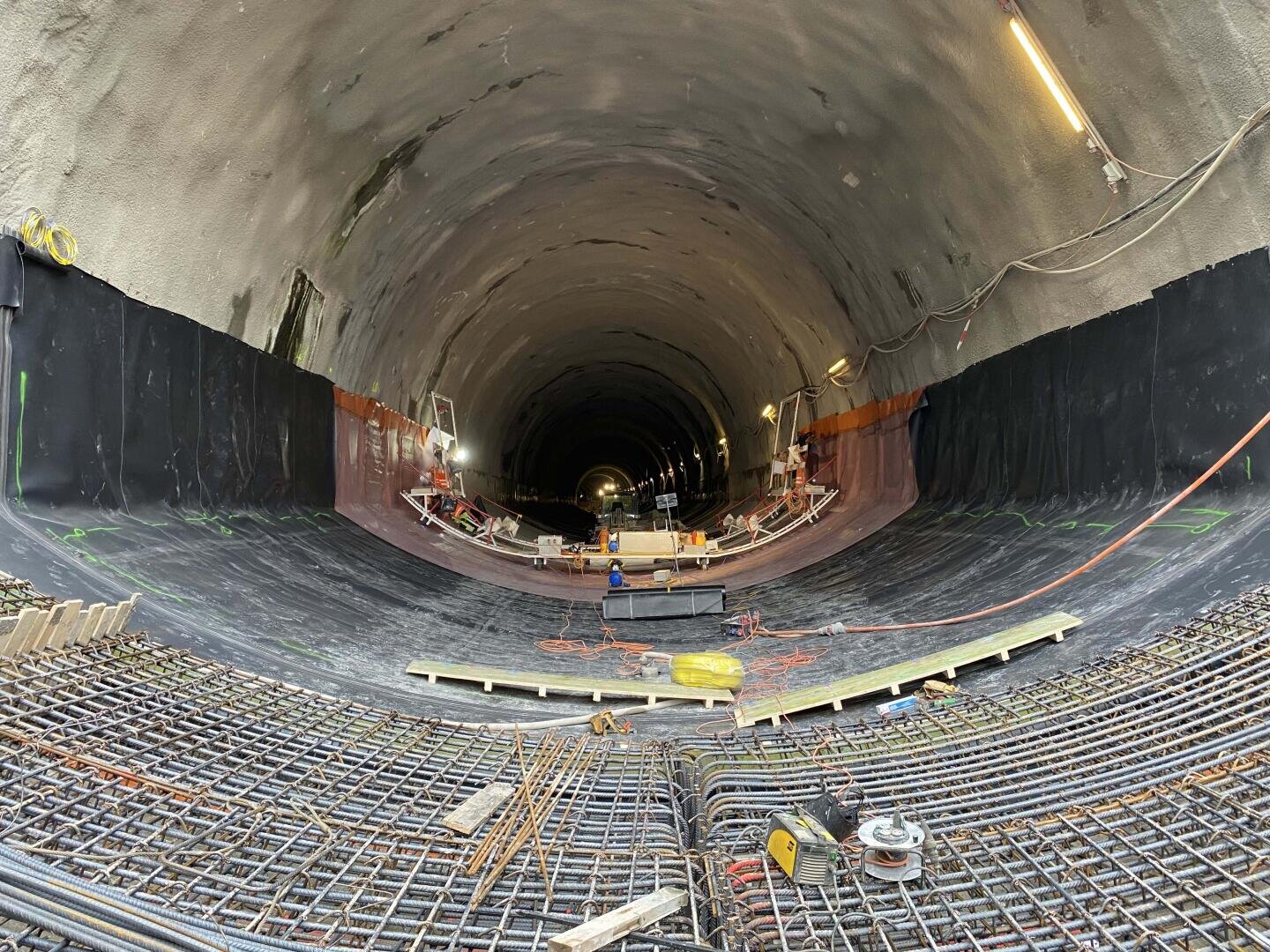 Ein großer, teilweise gebauter Tunnel mit freiliegendem Bewehrungsstahl und auf dem Boden verstreutem Baumaterial. Im Inneren sind Arbeiter und Geräte zu sehen. Der Tunnel wird durch an den Wänden angebrachte Oberlichter beleuchtet.