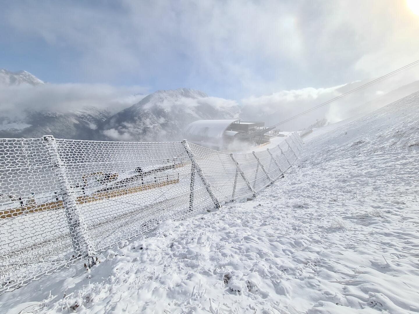 Eine schneebedeckte Skipiste mit einem Sicherheitszaun aus Metall, eine Skiliftstation im Hintergrund und neblige Berge unter einem teilweise bewölkten Himmel.