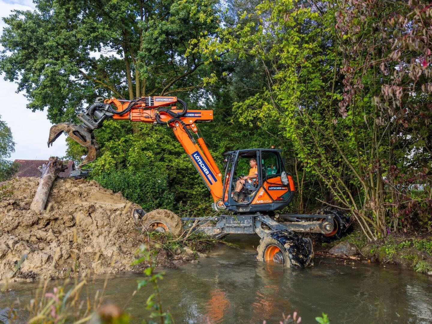 Ein orangefarbener Bagger mit einem Krallenaufsatz arbeitet am Rande eines schlammigen, flachen Baches und hebt ein großes Stück Holz an, das von grünen Bäumen und Sträuchern umgeben ist. Im Inneren der Kabine ist eine Person zu sehen.