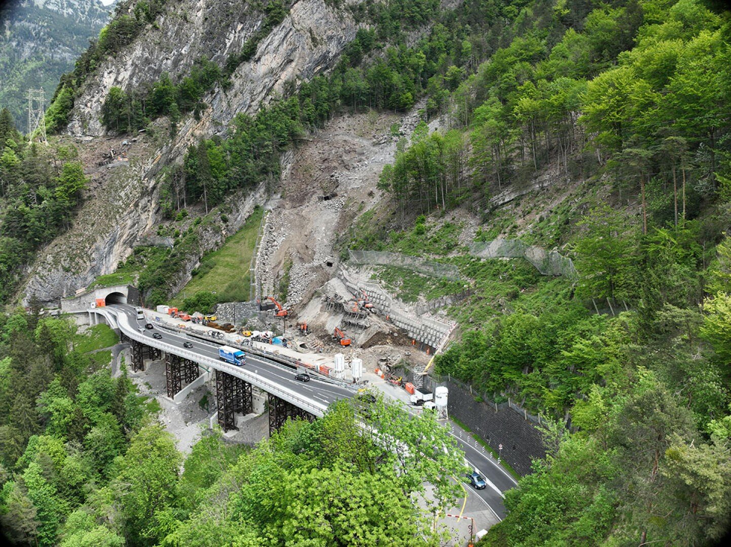 Luftaufnahme eines Erdrutsches an einem Berghang neben einem Autobahntunnel, mit Baufahrzeugen und Arbeitern, die die Trümmer beseitigen; üppig grüne Bäume und felsiges Gelände umgeben die Szene.