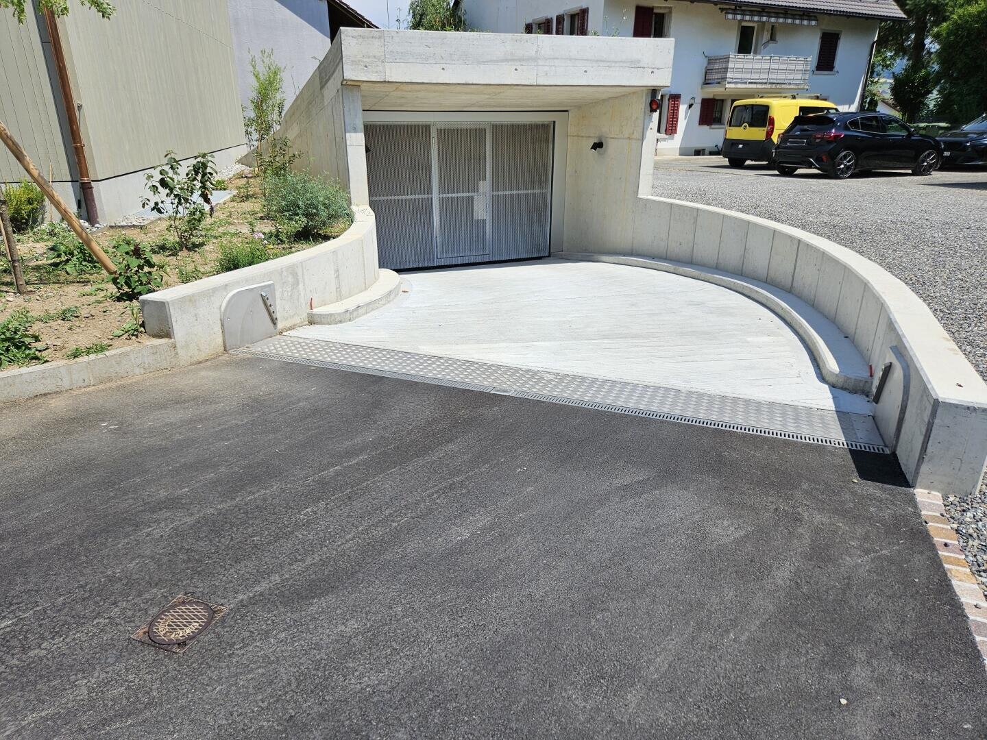 Eine moderne, geschwungene Einfahrt führt zu einem geschlossenen Garagentor, das in eine Betonwand eingelassen ist; in der Nähe befinden sich Pflanzen, ein Gehweg und geparkte Autos vor Wohnhäusern an einem sonnigen Tag.