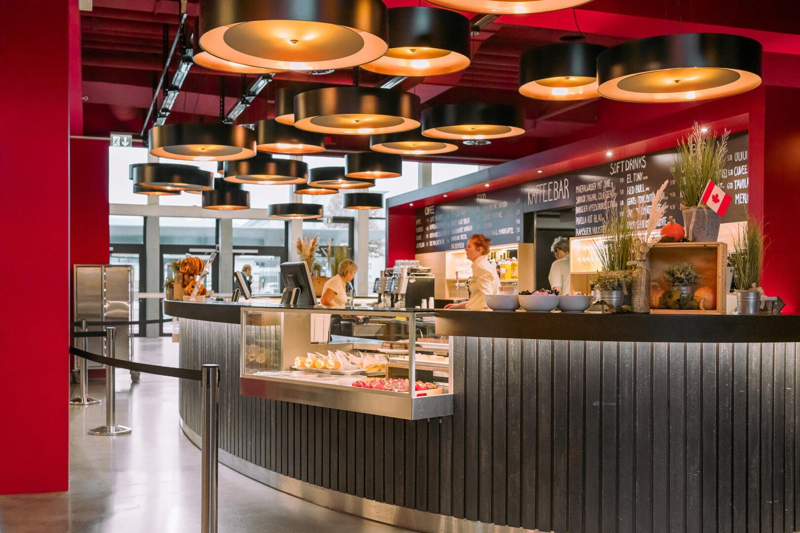 Eine moderne Cafeteria mit roten Wänden und runden Hängelampen. Das Personal hinter dem Tresen bereitet das Essen zu, während Gebäck und Getränke angeboten werden. Im Hintergrund sind Menütafeln zu sehen. Der Raum wirkt sauber und einladend.