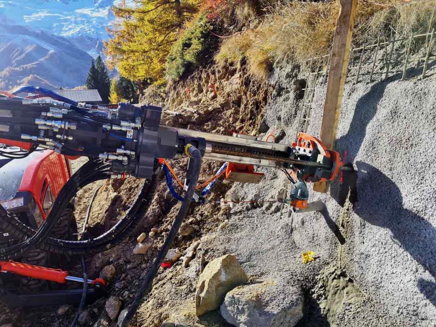 Eine Bohrmaschine ist auf felsigem Gelände montiert und bohrt Löcher in einen steilen, mit einem Metallgeflecht bewehrten Hang, während im Hintergrund Bäume und Berge zu sehen sind.