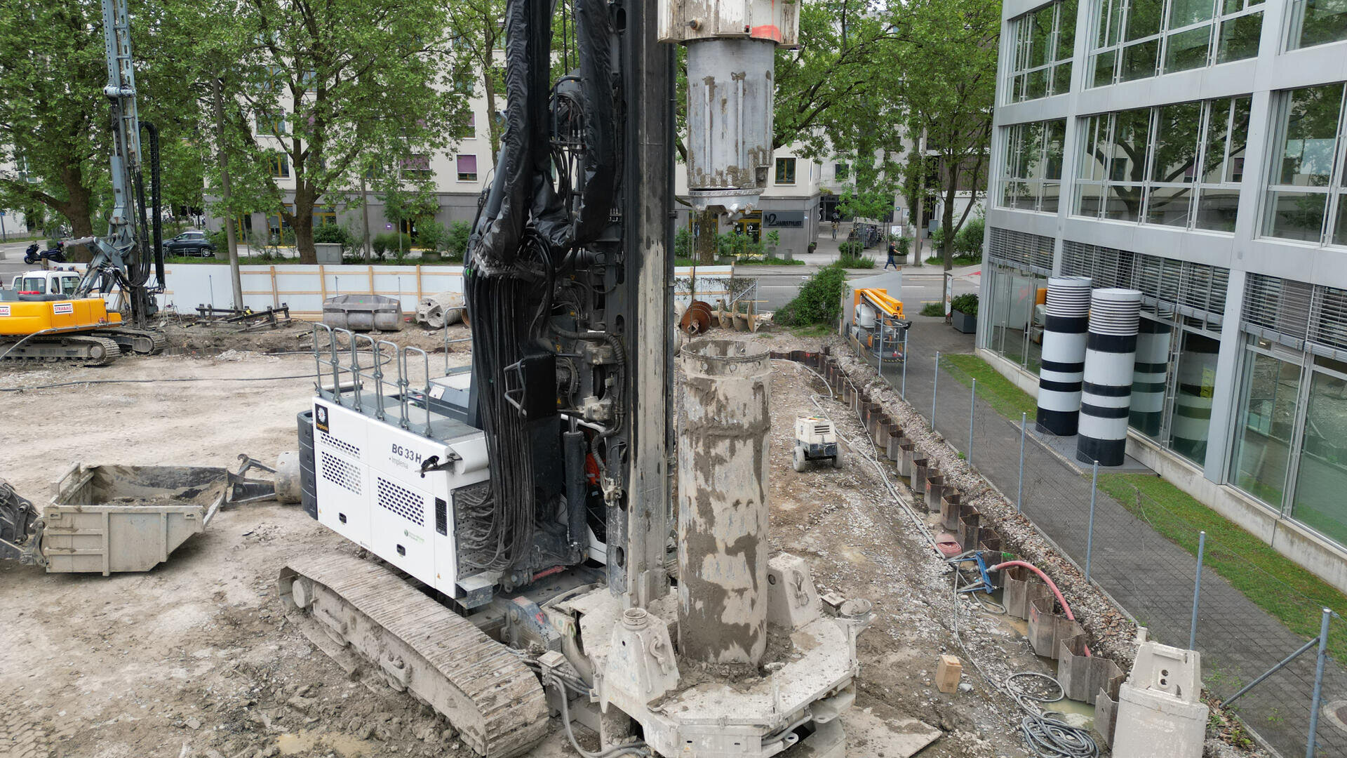 Eine große Bohrmaschine ist auf einer Baustelle in der Nähe eines modernen Glasgebäudes im Einsatz. Im Hintergrund sind weitere Baumaschinen, Rohre und Bäume zu sehen.