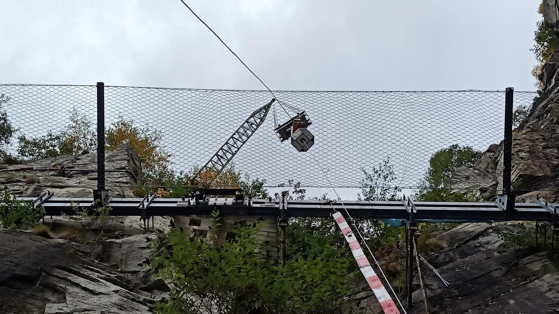 Eine Baustelle an einem felsigen Hang mit einem Metallzaun, einem Hängekran, der Material hebt, und einer Sicherheitsbarriere im Vordergrund. Spärliche Vegetation und bewölkter Himmel sind zu sehen.
