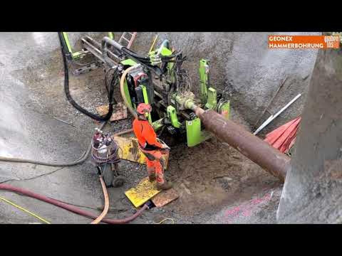 Ein Bauarbeiter in orangefarbener Schutzkleidung bedient schwere Maschinen, um auf einer schlammigen Baustelle in den Boden zu bohren, wobei Rohre und Geräte in der Umgebung zu sehen sind.