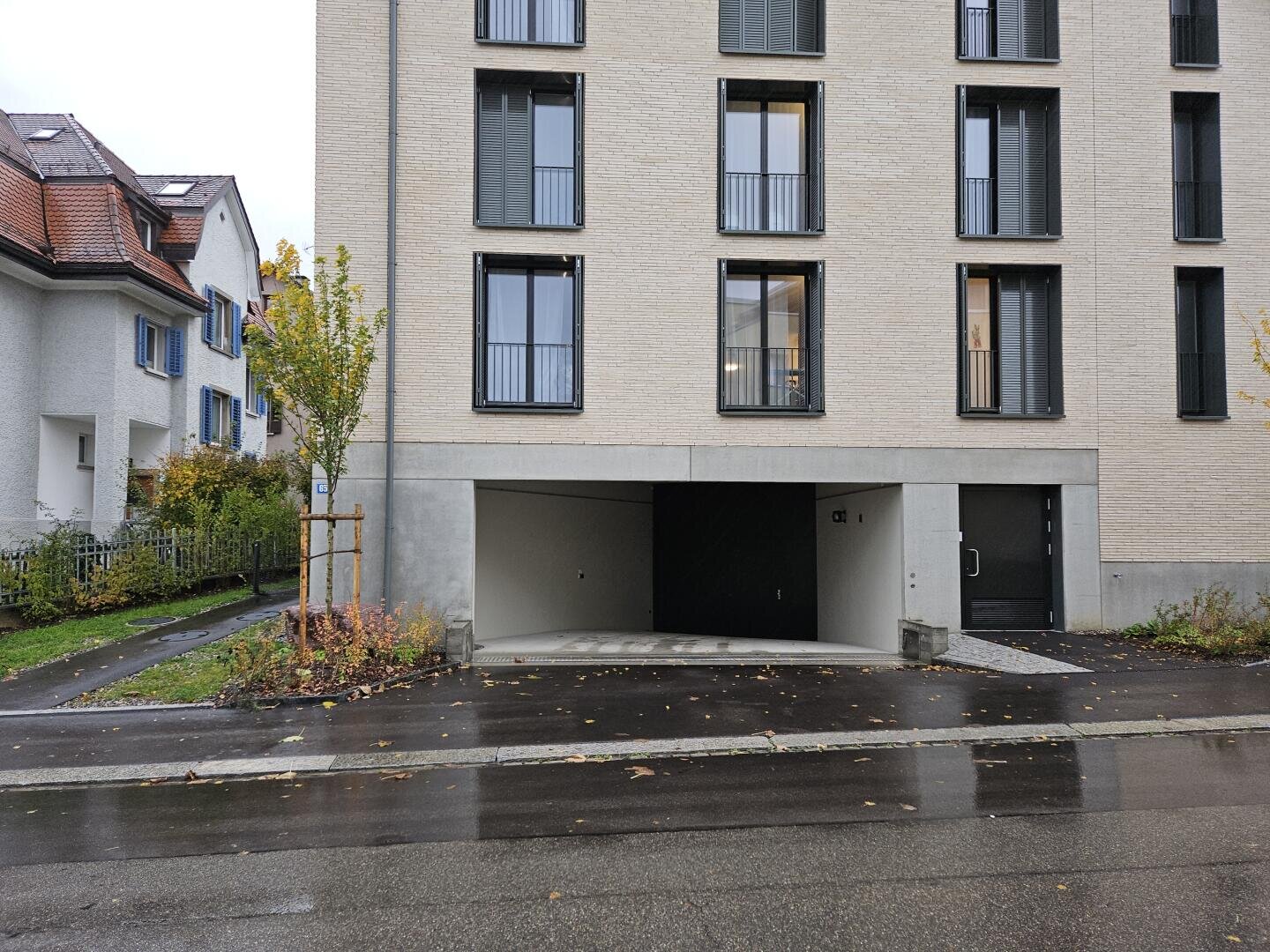 Ein modernes Mehrfamilienhaus mit hellbraunen Ziegeln verfügt über eine Einfahrt, die zu einer überdachten Parkgarage führt. An einem bewölkten Tag sind der nasse Bürgersteig, die Herbstbäume und die benachbarten Häuser zu sehen.