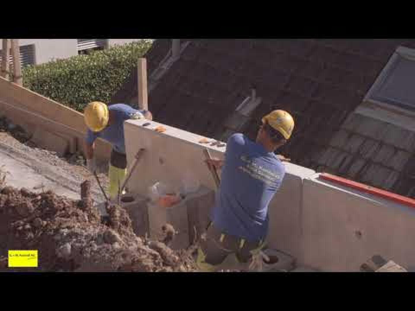 Zwei Bauarbeiter, die gelbe Helme und blaue Hemden tragen, bauen im Freien eine Betonwand. Werkzeuge und Materialien liegen in der Nähe verstreut, und es scheint sich um eine Baustelle in einem abschüssigen Gelände zu handeln.