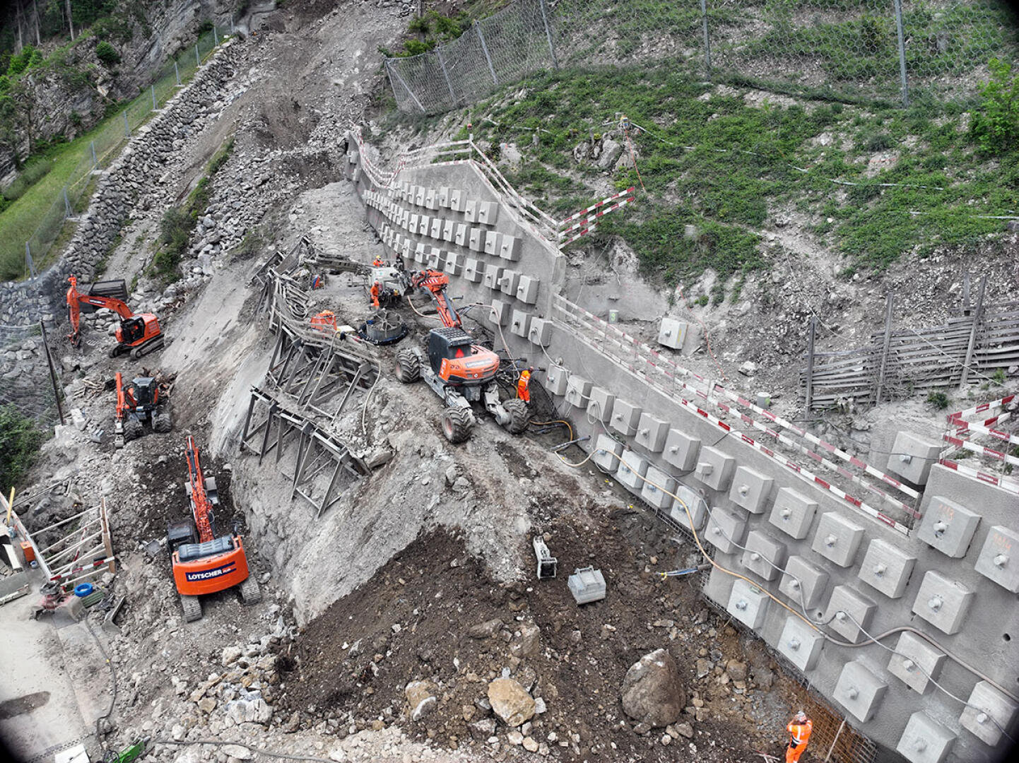 Luftaufnahme einer Baustelle an einem Hang mit Baggern, Arbeitern in orangefarbener Schutzkleidung, verstärkten Stützmauerblöcken, Erde, Steinen und Zäunen entlang des Hangs.