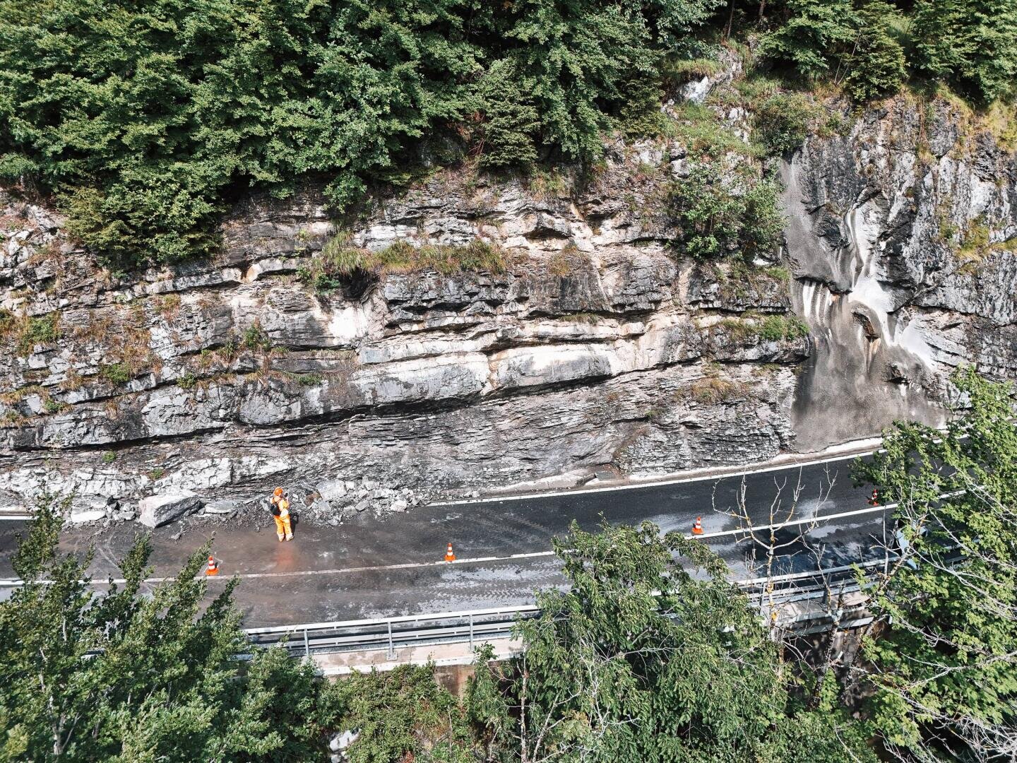 Ein Arbeiter in orangefarbener Schutzkleidung steht auf einer kurvenreichen Bergstraße neben einer felsigen Klippe, mit Verkehrsleitkegeln am Straßenrand und dichten grünen Bäumen über ihm.
