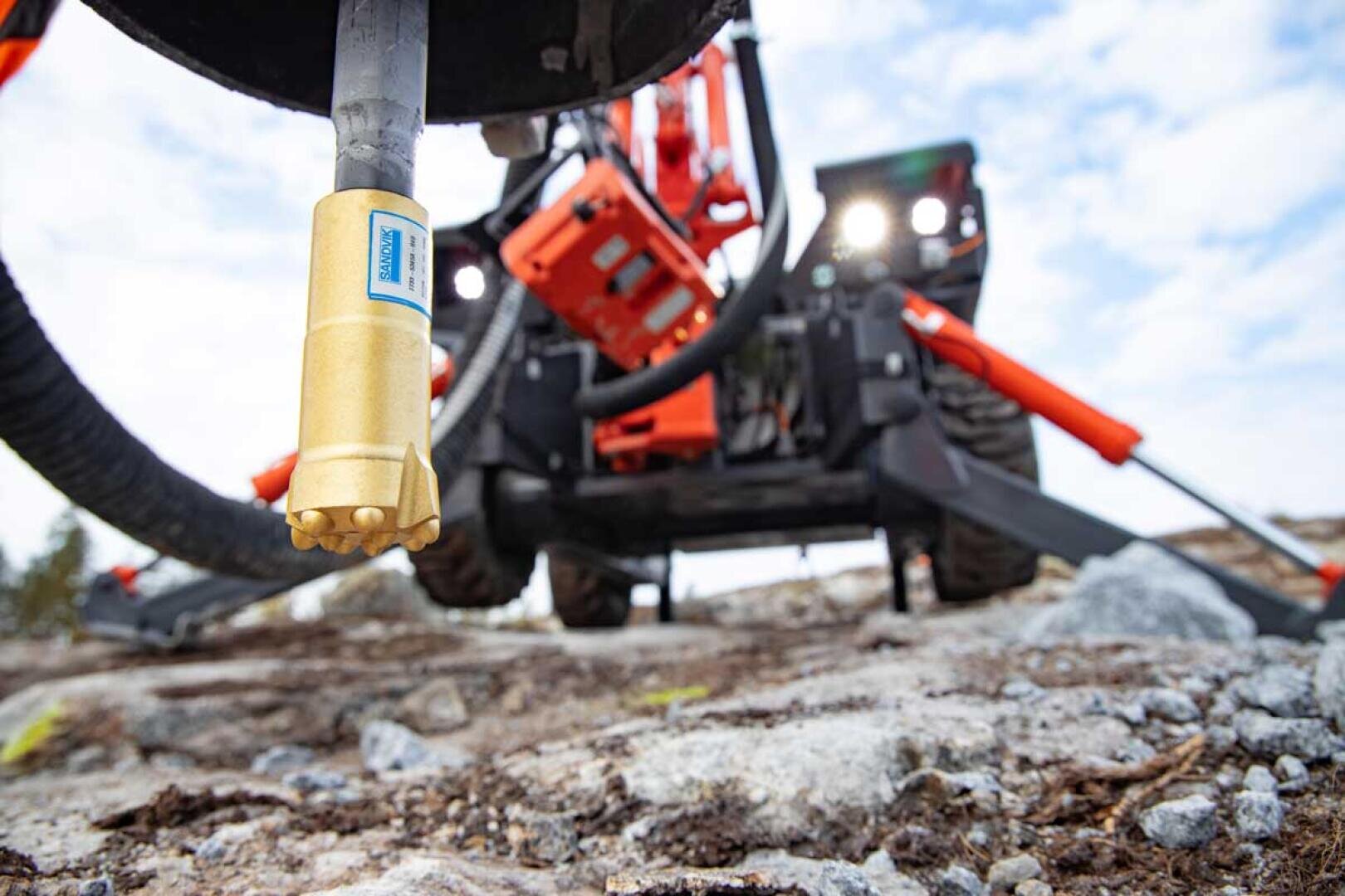 Nahaufnahme eines Goldbohrers, der an einer schweren Maschine befestigt ist, wobei die Maschine und das felsige Gelände im Hintergrund verschwimmen. Der Schwerpunkt liegt auf dem Bohrer, der die technische Ausrüstung für den Bergbau oder das Bauwesen hervorhebt.
