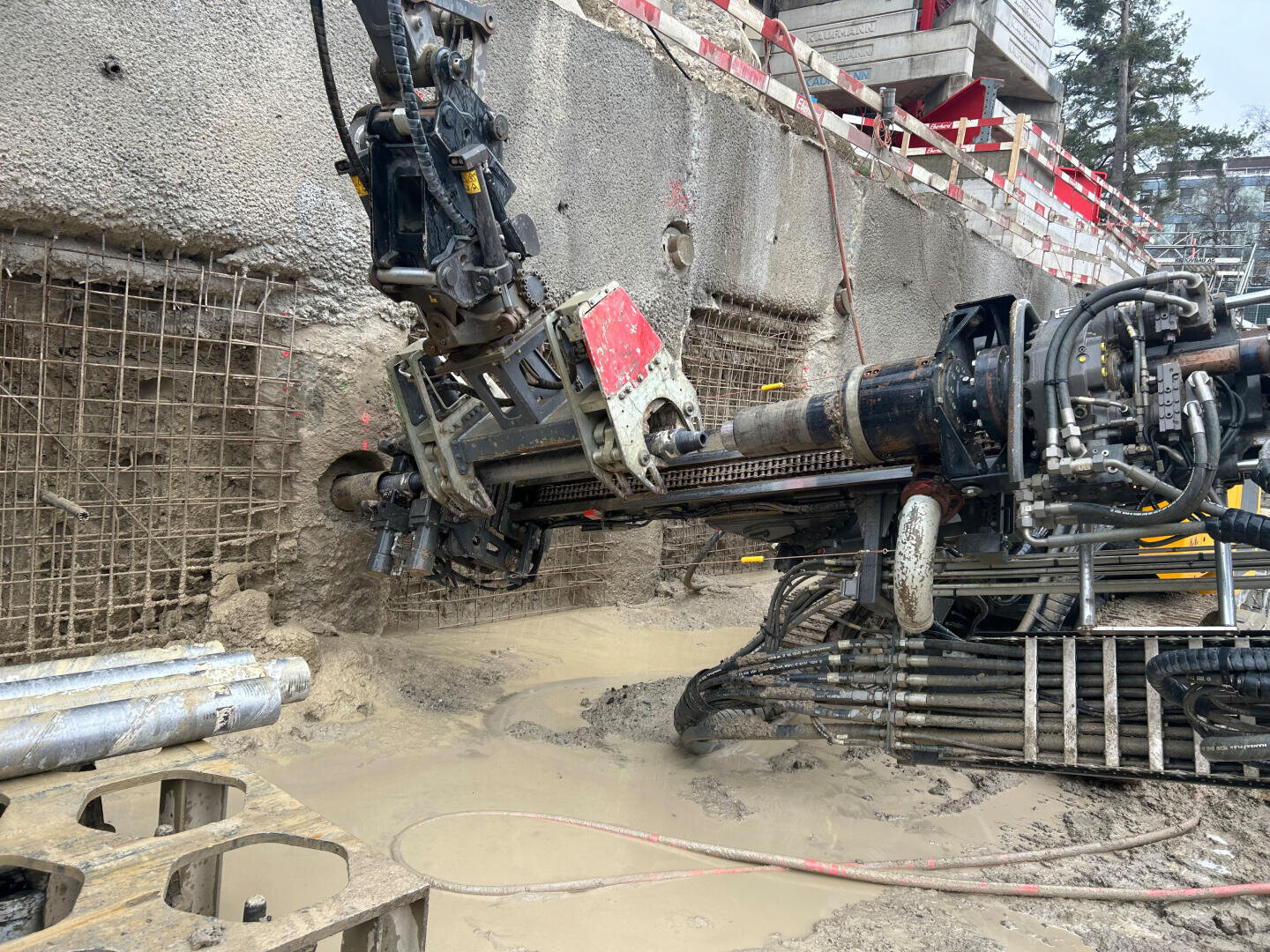 Eine große Horizontalbohrmaschine bohrt sich auf einer schlammigen Baustelle in eine Wand aus Beton und Armierungseisen, um die herum Kabel, Rohre und Bauabsperrungen zu sehen sind.