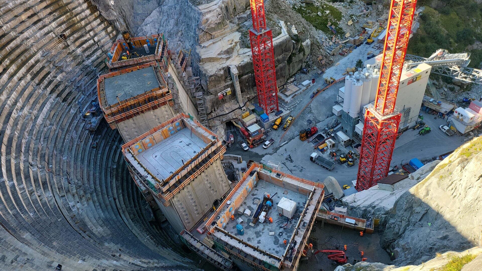 Luftaufnahme einer großen Baustelle in einer tiefen, kreisförmigen Grube mit drei hohen, teilweise errichteten Betonstrukturen und roten Kränen, umgeben von Maschinen und Arbeitern.
