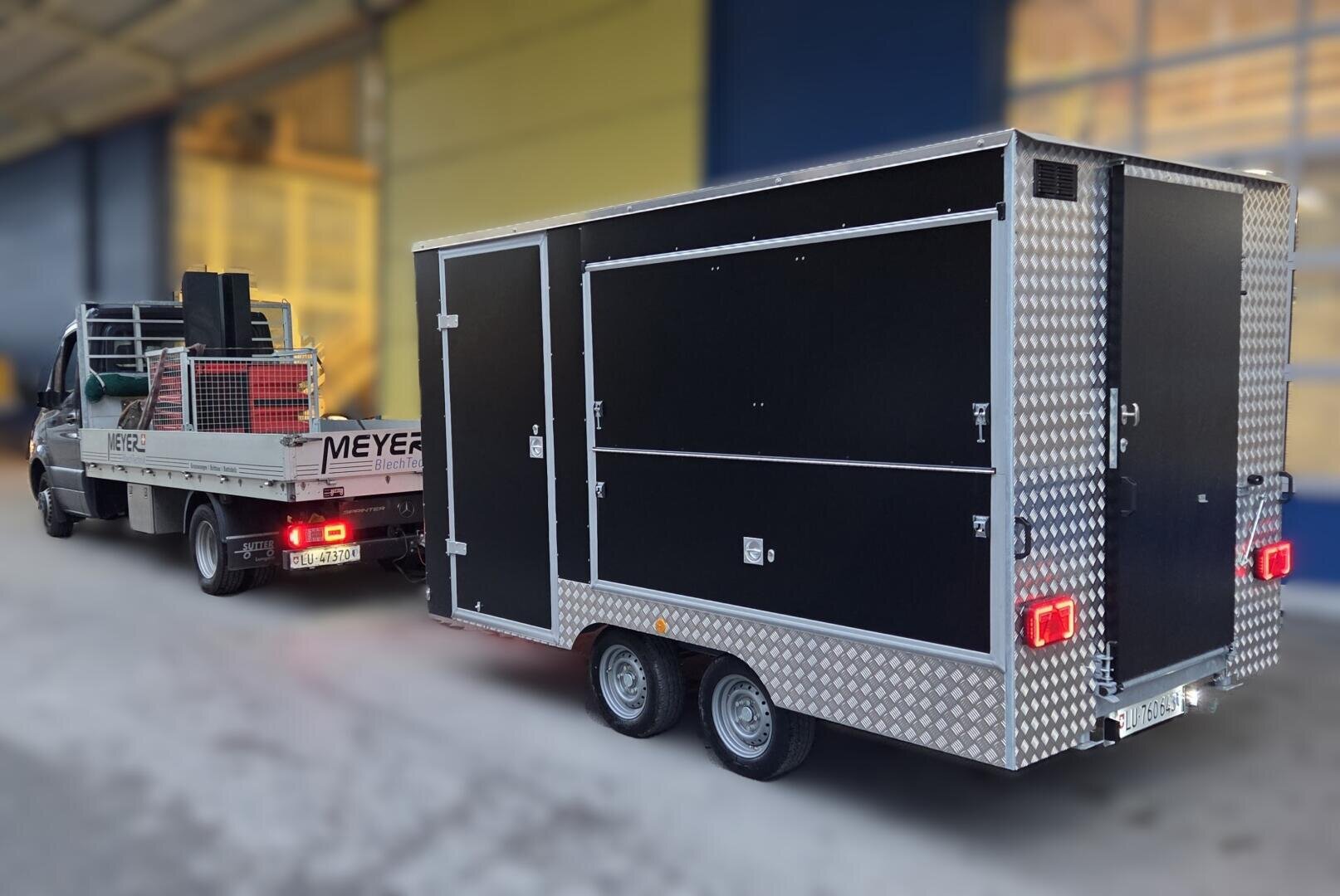 Ein Pritschenwagen mit MEYER-Schriftzug zieht vor einem Industriegebäude einen großen, zweiachsigen, schwarz-silbernen Nutzfahrzeuganhänger. Der Anhänger ist mit mehreren Fächern und Rückleuchten ausgestattet.