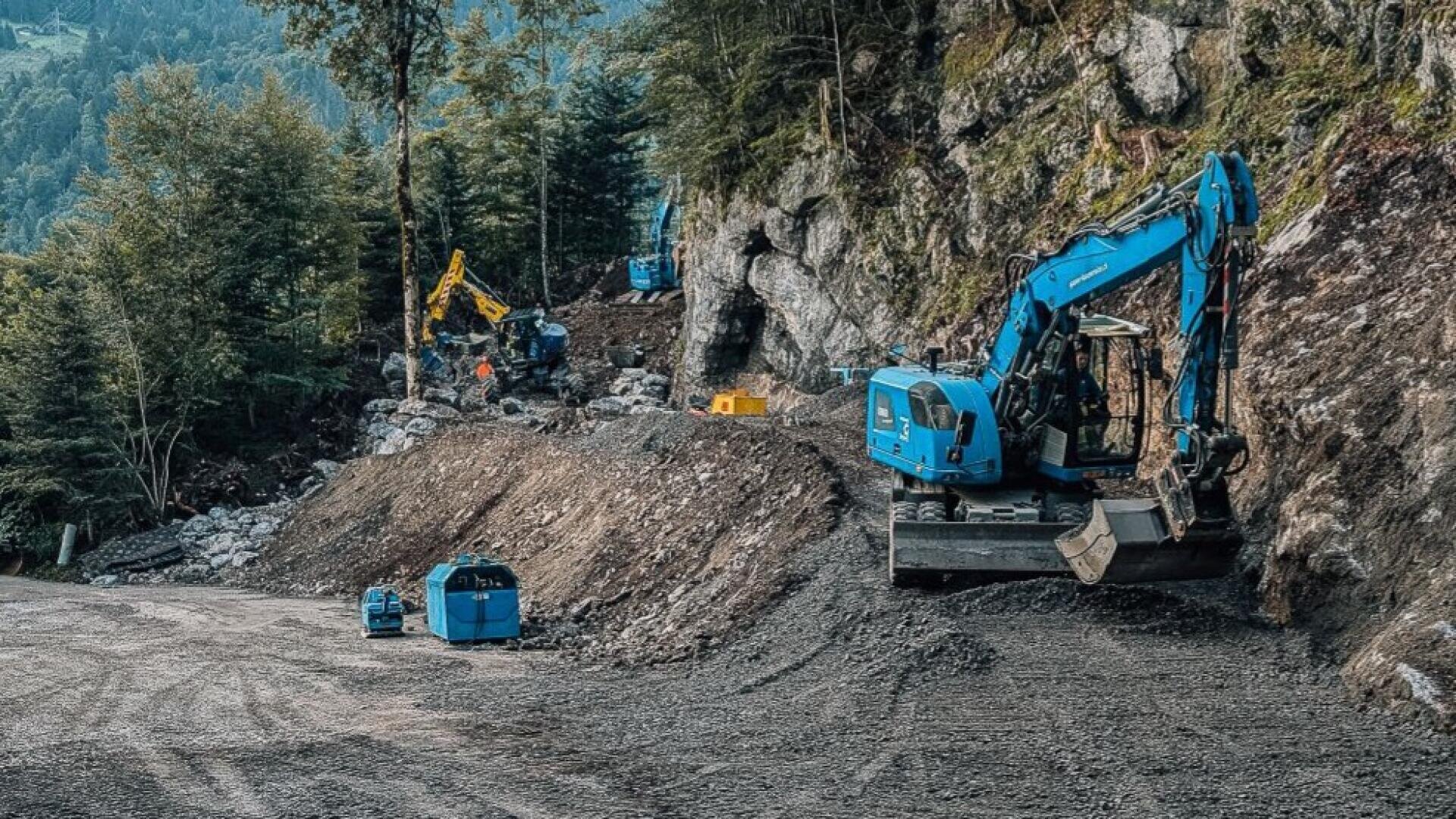 Mehrere blaue Bagger und Bauarbeiter sind dabei, einen felsigen, bewaldeten Hang zu roden und einzuebnen, um eine Straße zu bauen. Bäume und Grünflächen umgeben die Baustelle, und steile Felswände säumen das Gelände.