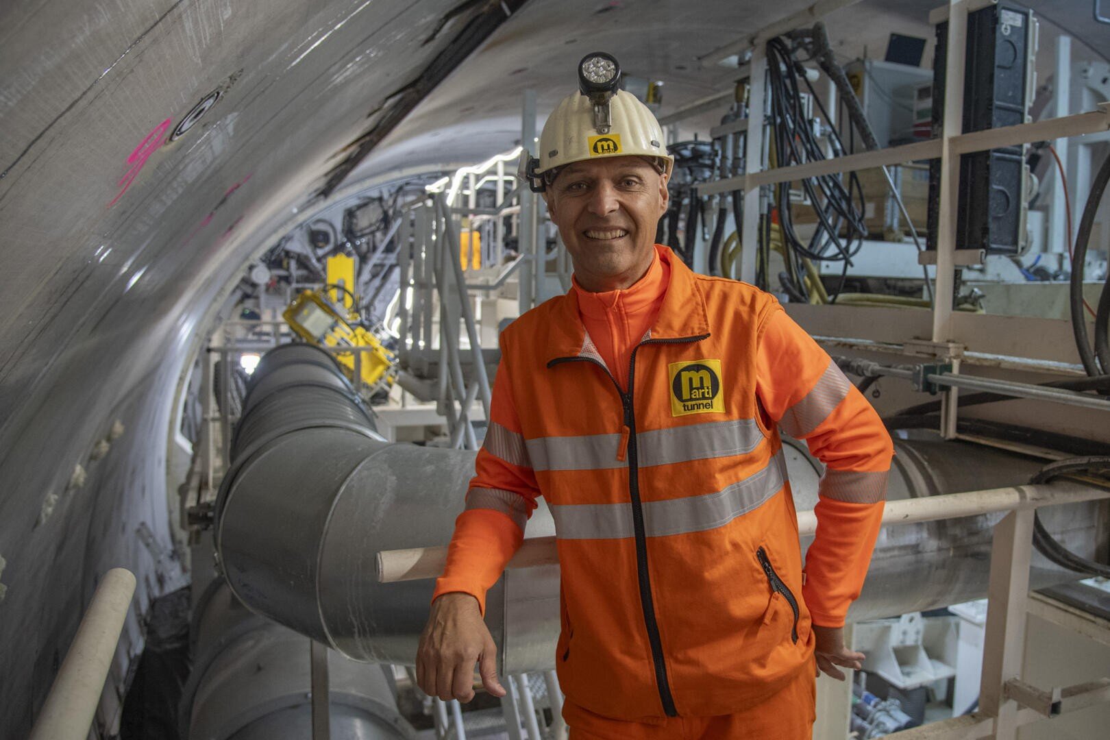 Ein Arbeiter in orangefarbenem Warnschutzanzug und weißem Schutzhelm steht in einem großen, im Bau befindlichen Tunnel, umgeben von Maschinen, Rohren und Gerüsten.