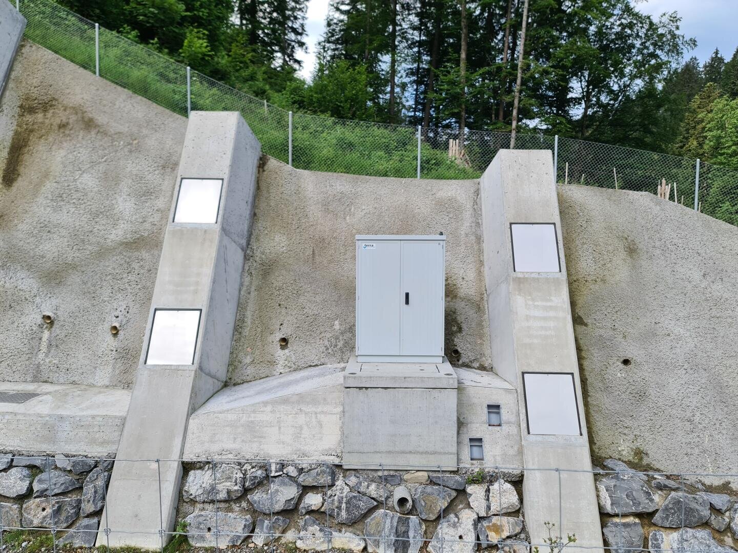Ein grauer Geräteschrank ist auf einer Betonplattform installiert, die in einen abfallenden Hang gebaut wurde, mit Betonstützmauern und einem Metallzaun darüber, umgeben von Bäumen und Grünflächen.