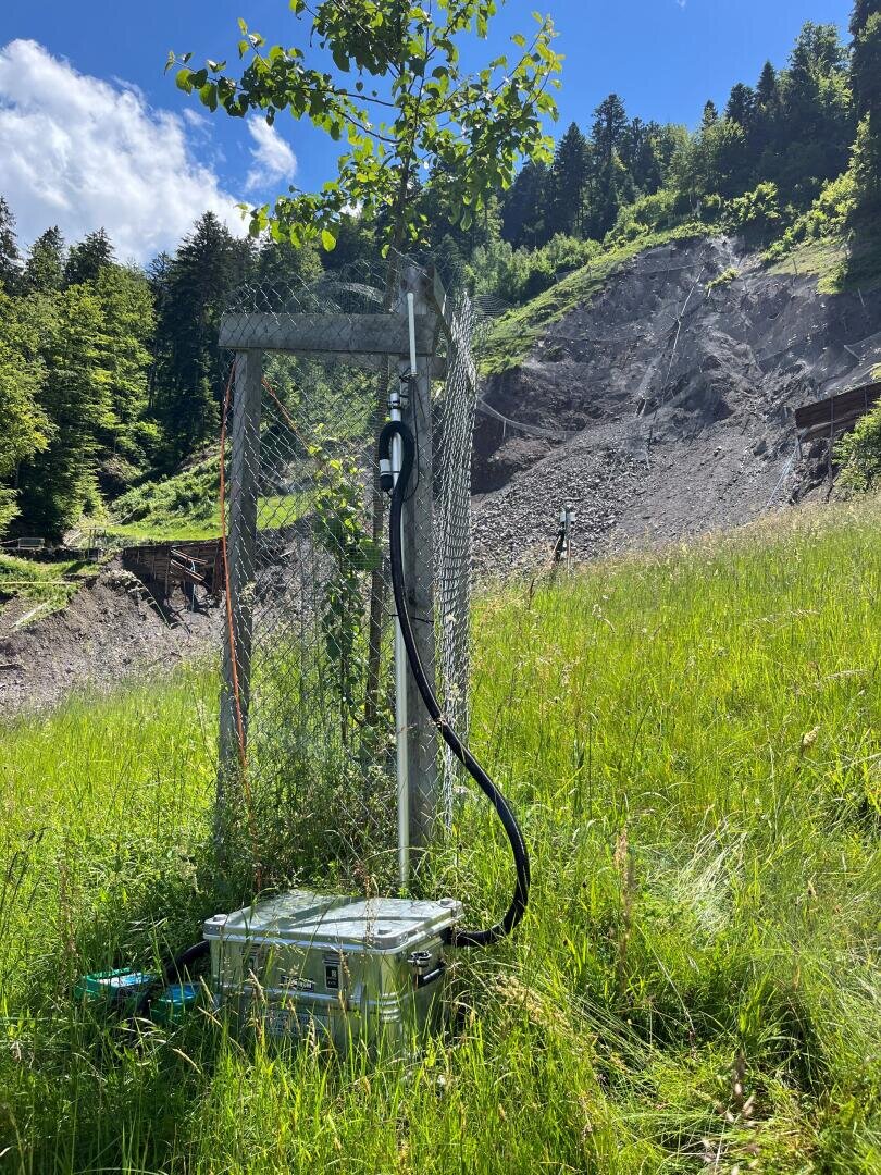 Ein junger Baum, umgeben von einem Drahtzaun, steht auf einem grasbewachsenen Hügel und ist über ein schwarzes Kabel mit einer Überwachungsanlage verbunden. Dahinter durchschneidet ein steiler, felsiger Erdrutsch den grünen Wald unter einem teilweise bewölkten Himmel.