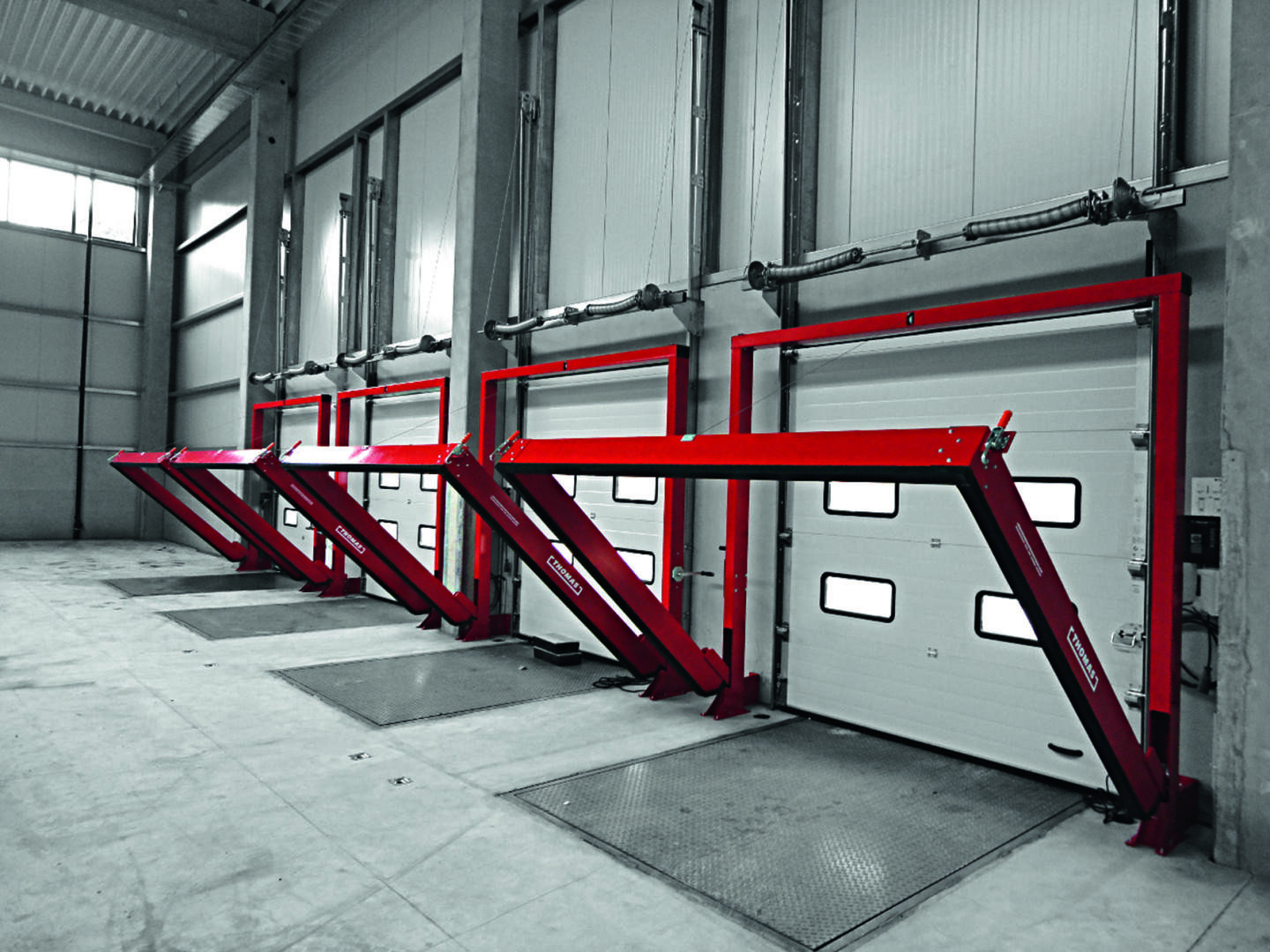 Vier rote Fahrzeughebebühnenarme sind vor geschlossenen Garagentoren in einer geräumigen, industriell anmutenden Anlage mit Betonböden und -wänden aufgestellt. Die Szene wirkt sauber und geordnet.