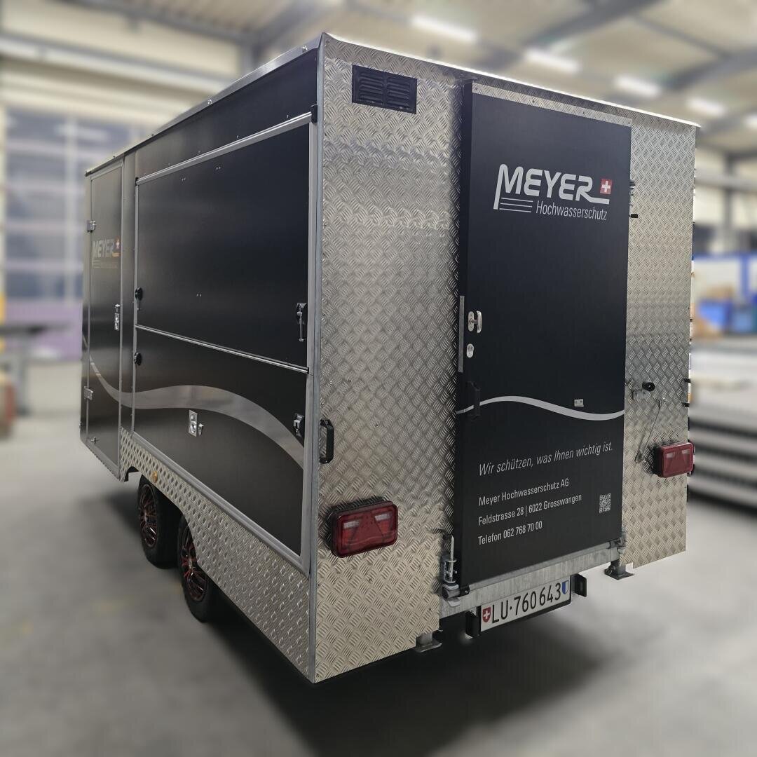 Ein silber-schwarzer, geschlossener Anhänger mit der Aufschrift MEYER Hochwasserschutz ist im Innenbereich geparkt. Der Anhänger hat eine Rautenverkleidung, ein Schweizer Nummernschild und Kontaktinformationen auf der Hintertür.