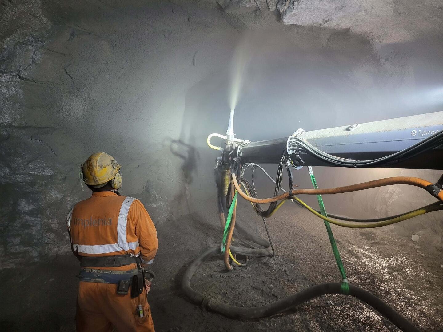 Ein Arbeiter in orangefarbener Schutzkleidung und mit Helm bedient schwere Maschinen, die Wasser oder Beton auf die Felswand in einem unterirdischen Tunnel spritzen. Schläuche und Kabel sind mit dem Gerät verbunden.
