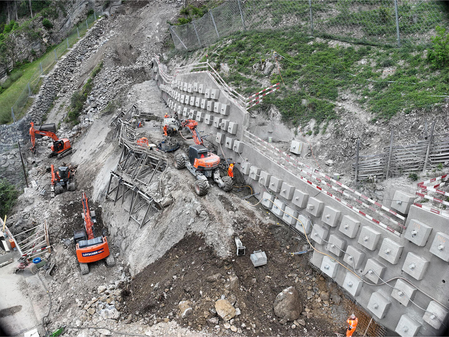 Eine Baustelle an einem steilen Hang mit mehreren orangefarbenen Baggern, Betonbarrieren und Arbeitern in orangefarbener Schutzkleidung, die den Hang in der Nähe eines Zauns und felsigen Geländes stabilisieren.