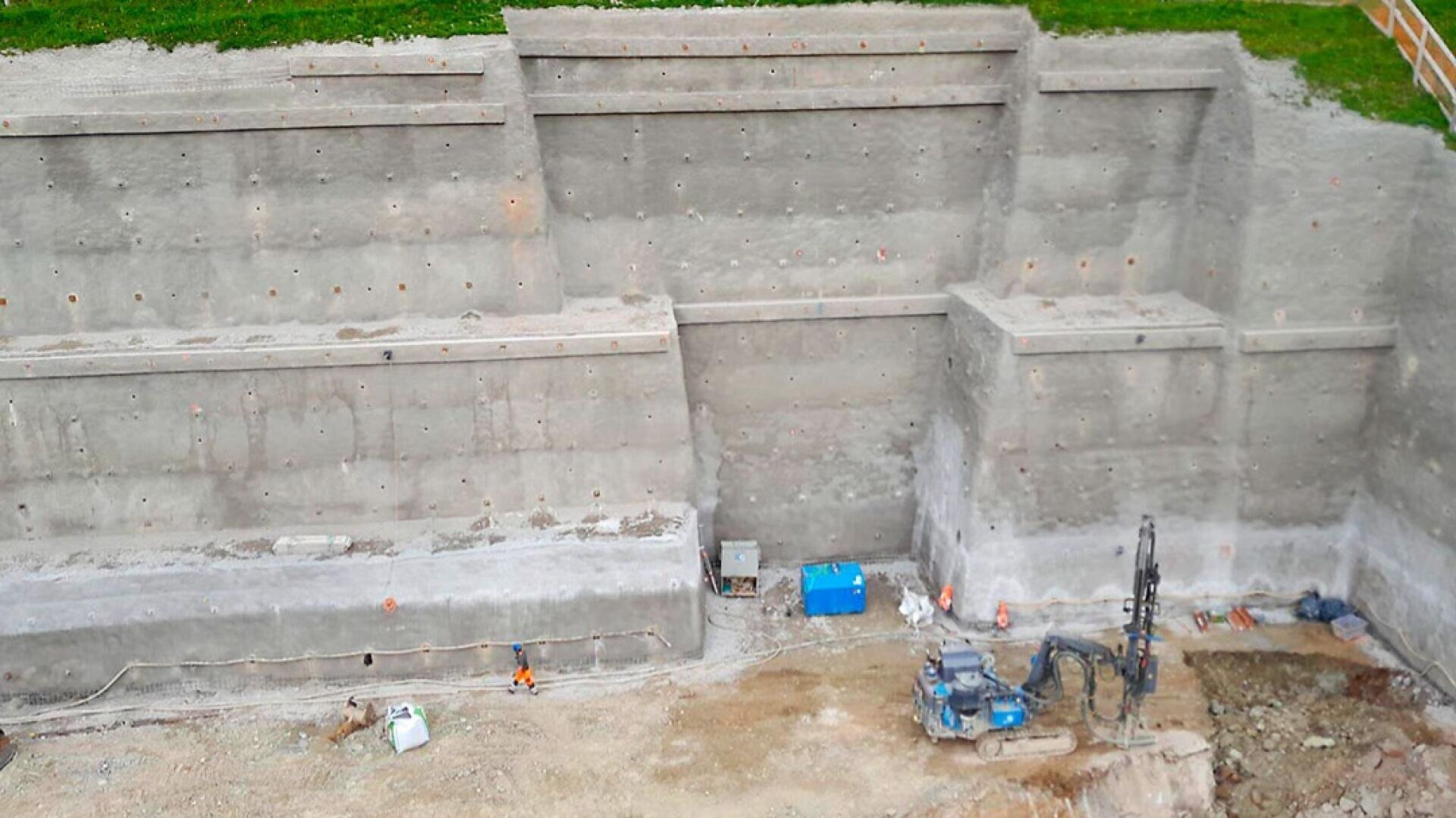 Eine Stahlbetonstützmauer mit mehreren abgestuften Ebenen ist in einen Erdhang gebaut. In der Nähe der Mauer liegen Baugeräte, Kabel und Material auf dem Boden, und am oberen Rand ist Gras zu sehen.