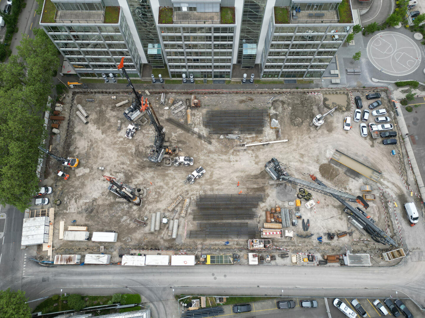 Luftaufnahme einer Baustelle mit Kränen und Maschinen, umgeben von Straßen, geparkten Autos und modernen Gebäuden. Auf der Baustelle sind rechteckige Fundamente und Baumaterialien zu sehen.