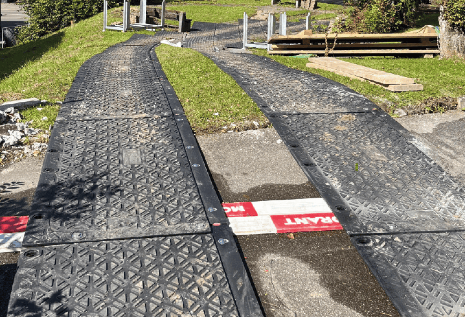Zwei schwarze Kunststoffmatten bilden provisorische Wege über Gras und Pflaster, umgeben von Baumaterialien und -geräten, wobei der helle Sonnenhimmel klare Schatten wirft.
