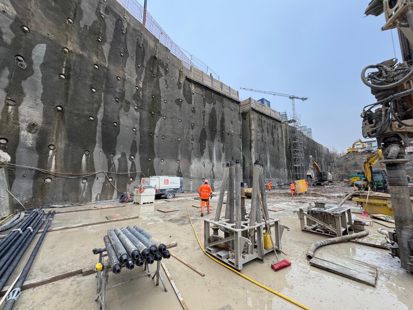 Baustelle mit tiefem Aushub, großer Stützmauer auf der linken Seite, Arbeitern in orangefarbener Schutzkleidung, Maschinen, Metallstangen und nassem, schlammigem Boden. Im Hintergrund sind Kräne und Gebäudestrukturen unter einem bewölkten Himmel zu sehen.