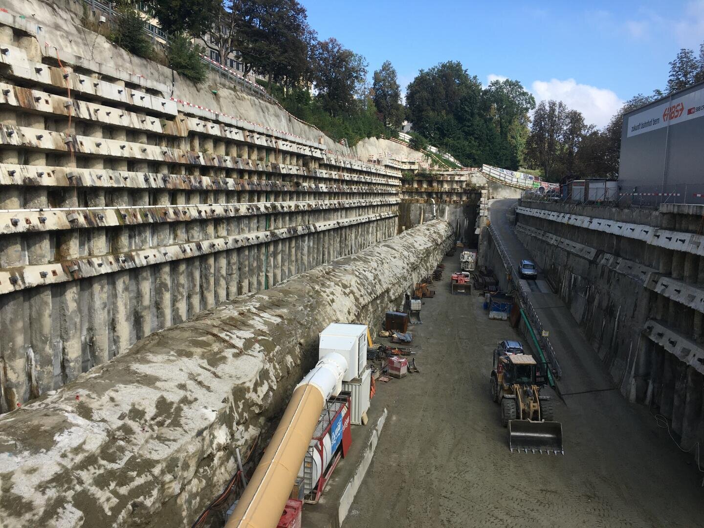 Eine große Baustelle für einen unterirdischen Tunnel, mit Betonstützmauern, schweren Maschinen und Geräten entlang eines unbefestigten Weges. Das Gelände ist von Bäumen und Gebäuden an den oberen Rändern umgeben.