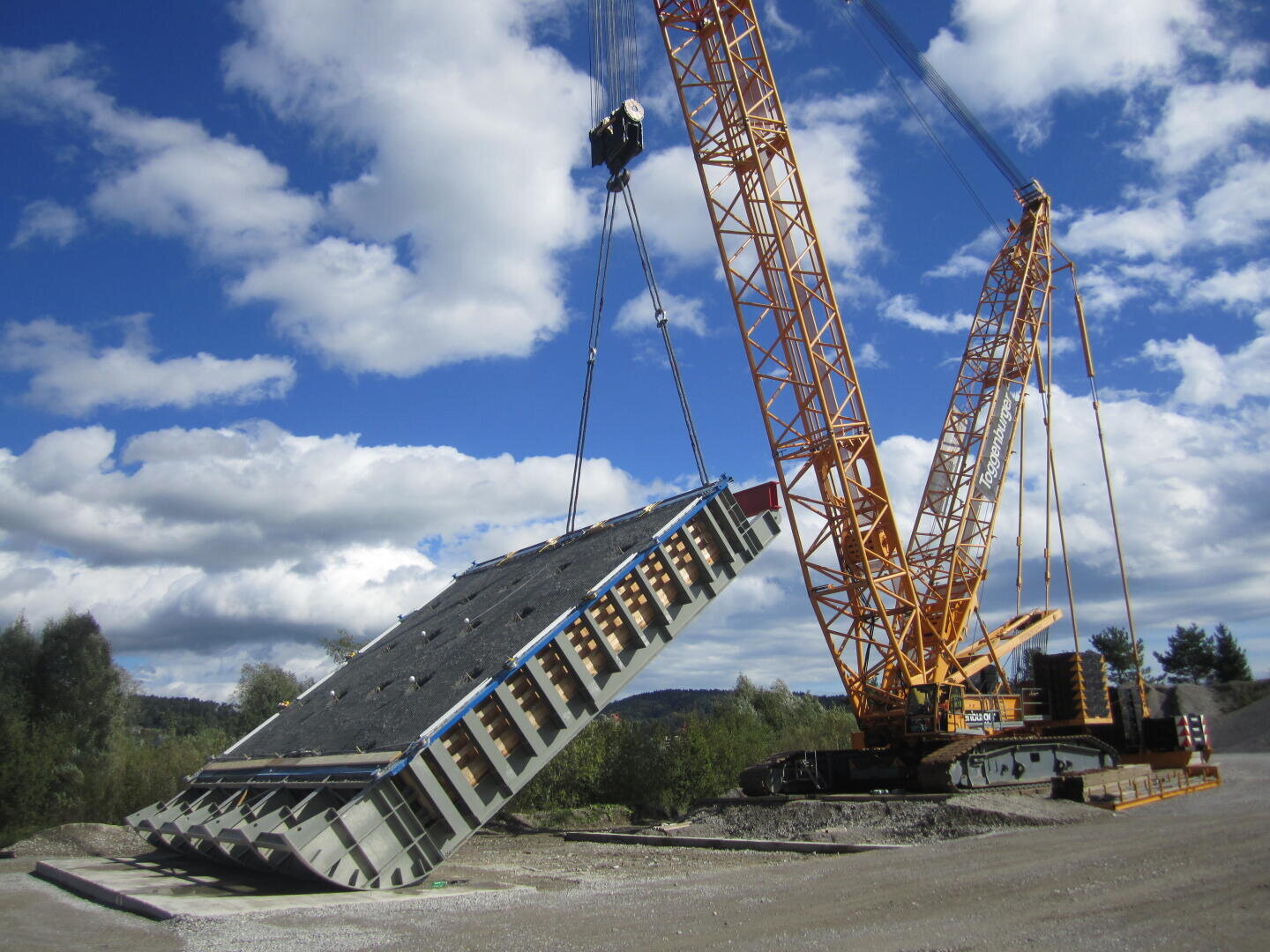 Ein großer gelber Kran hebt eine massive rechteckige Stahl- und Betonkonstruktion auf einer Baustelle unter einem teilweise bewölkten blauen Himmel an. Im Hintergrund sind Bäume und Schotter zu sehen.