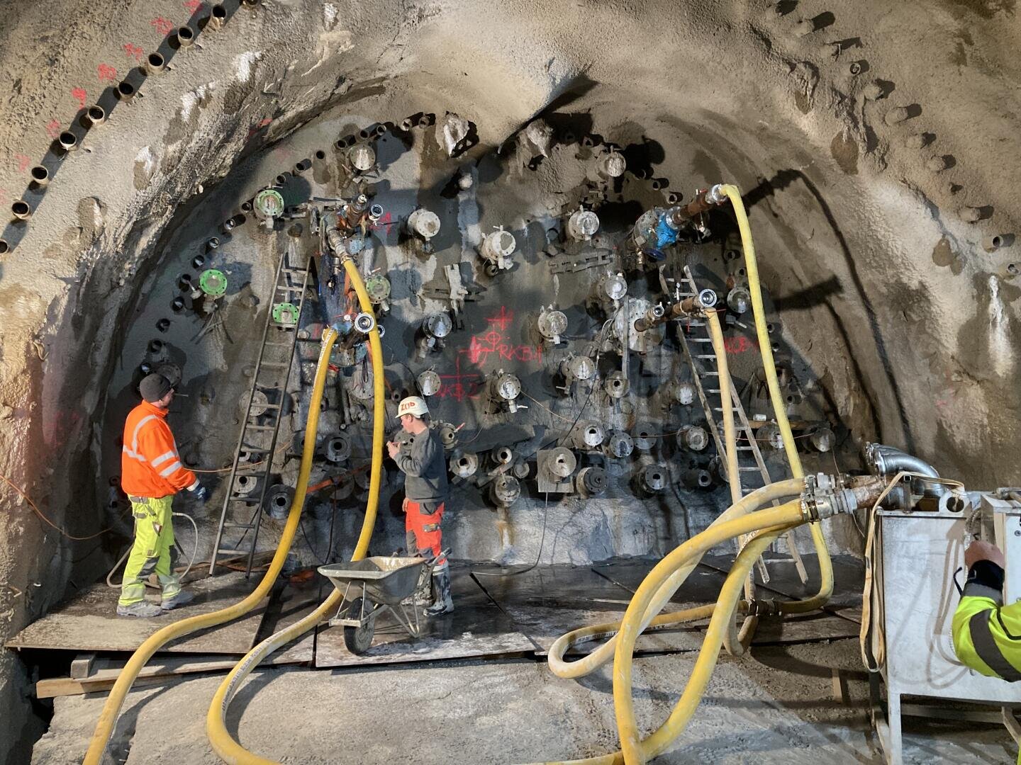 Drei Arbeiter in Schutzausrüstung bedienen Maschinen und Schläuche in einem im Bau befindlichen Tunnel. An der Tunnelwand sind mehrere kreisförmige Geräte angebracht, die durch Kabel und Schläuche miteinander verbunden sind, und an der Wand lehnen Leitern.