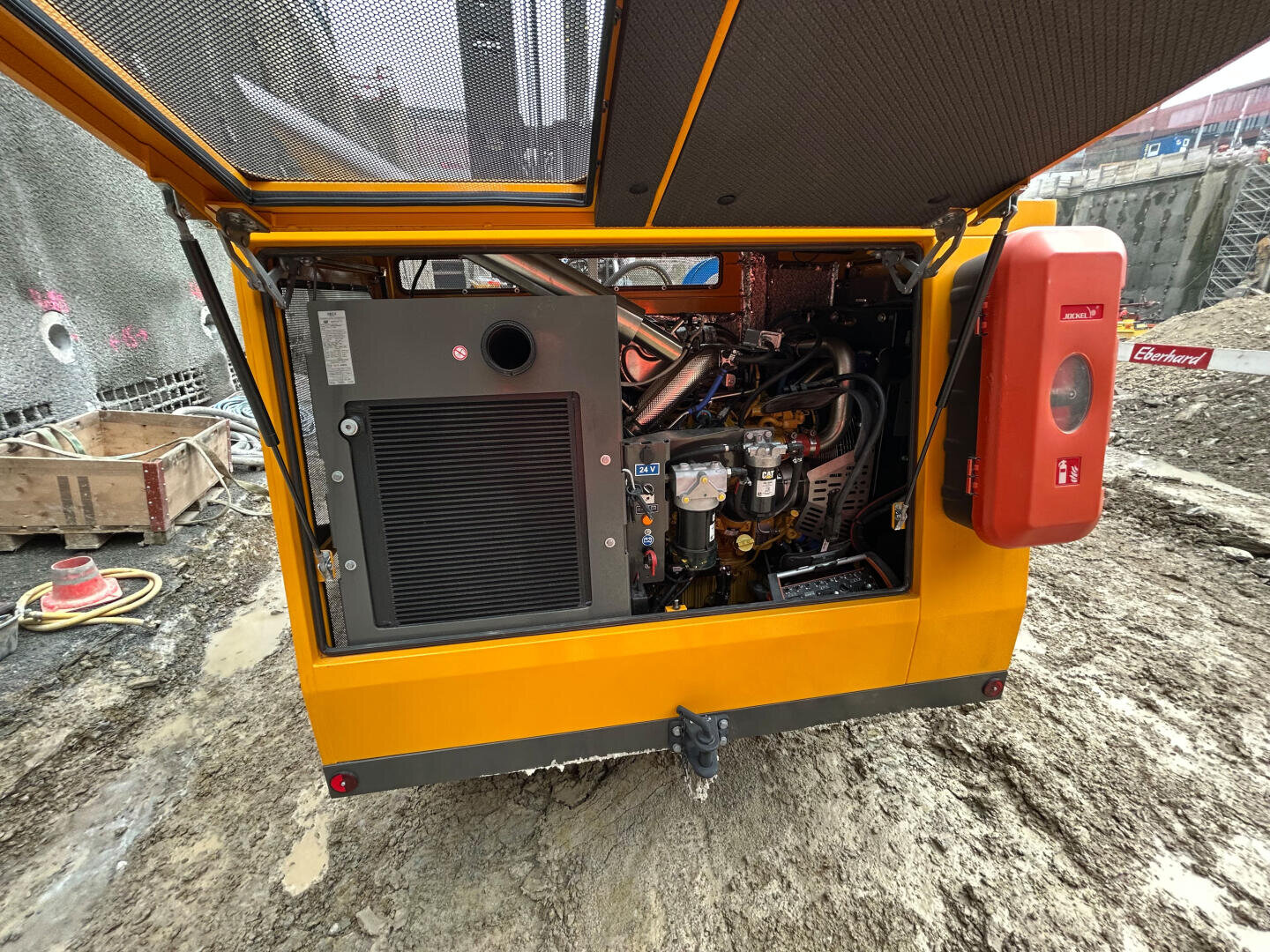 Nahaufnahme des offenen hinteren Fachs eines gelben mobilen Generators auf einer Baustelle, mit den Motorkomponenten und einem roten Notfallset an der Seite. In der Nähe sind schlammiger Boden und Baumaterialien zu sehen.