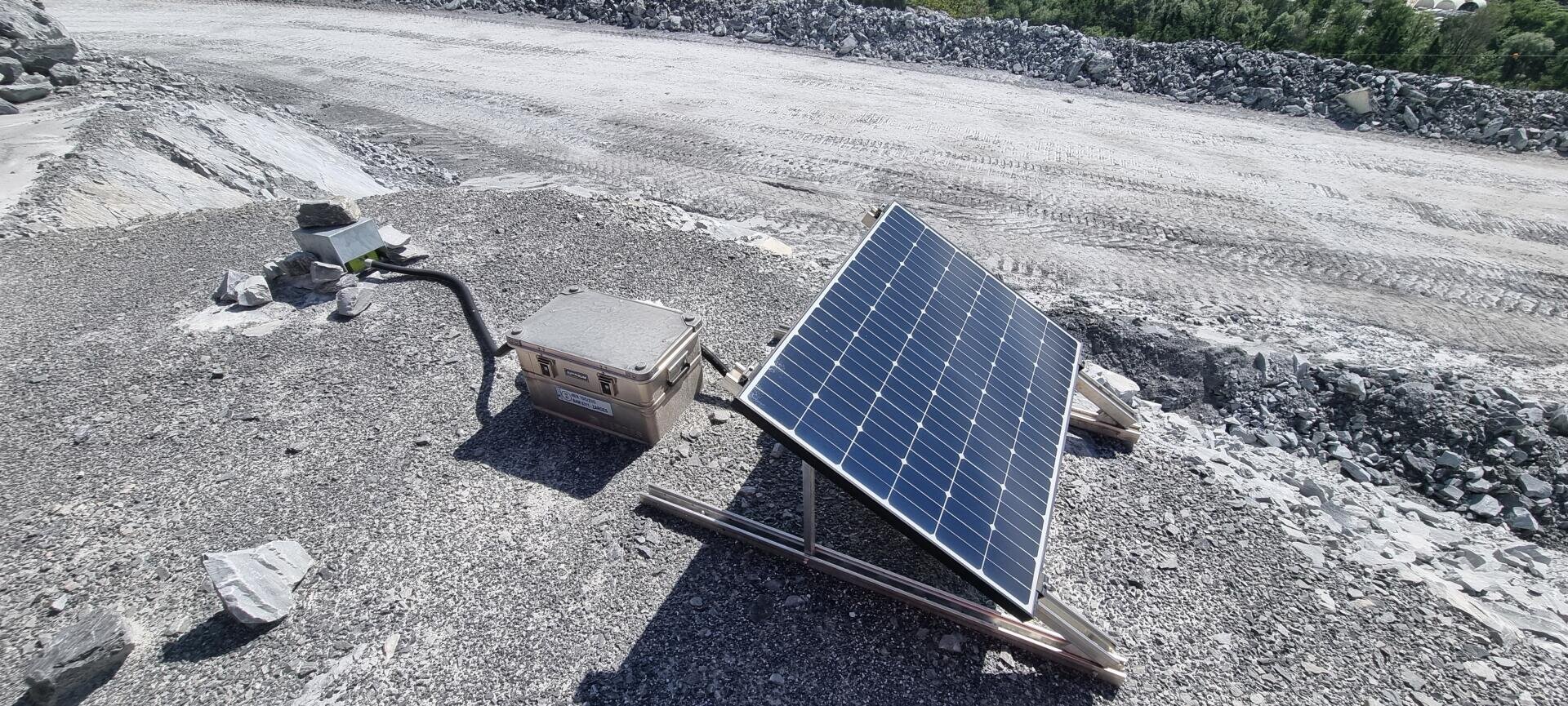 Ein Solarmodul ist auf einem Gestell montiert und mit einem beigefarbenen tragbaren Stromaggregat verbunden, das auf einer Schotterfläche auf einer Baustelle oder im Bergbau steht, umgeben von Felsen und Reifenspuren.
