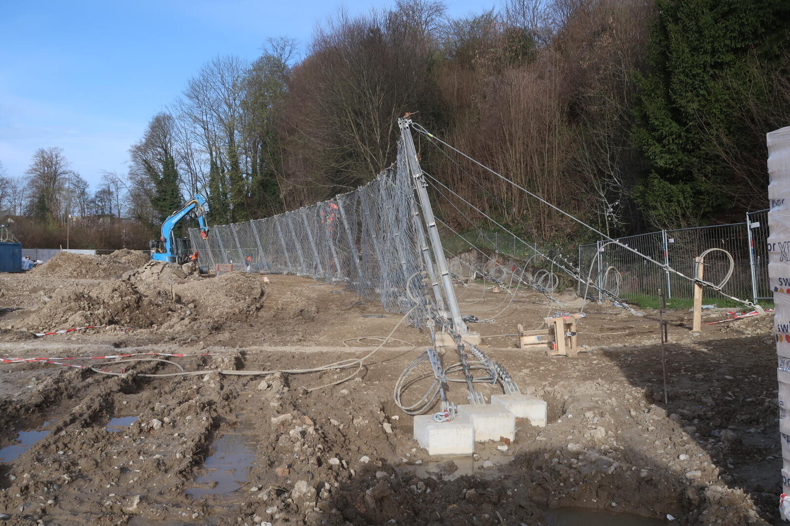 Eine Baustelle mit schlammigem Boden, einem gekippten Metallzaun, der von Kabeln gestützt wird, und einem blauen Bagger im Hintergrund. An einem klaren Tag begrenzen blattlose Bäume das Gebiet.